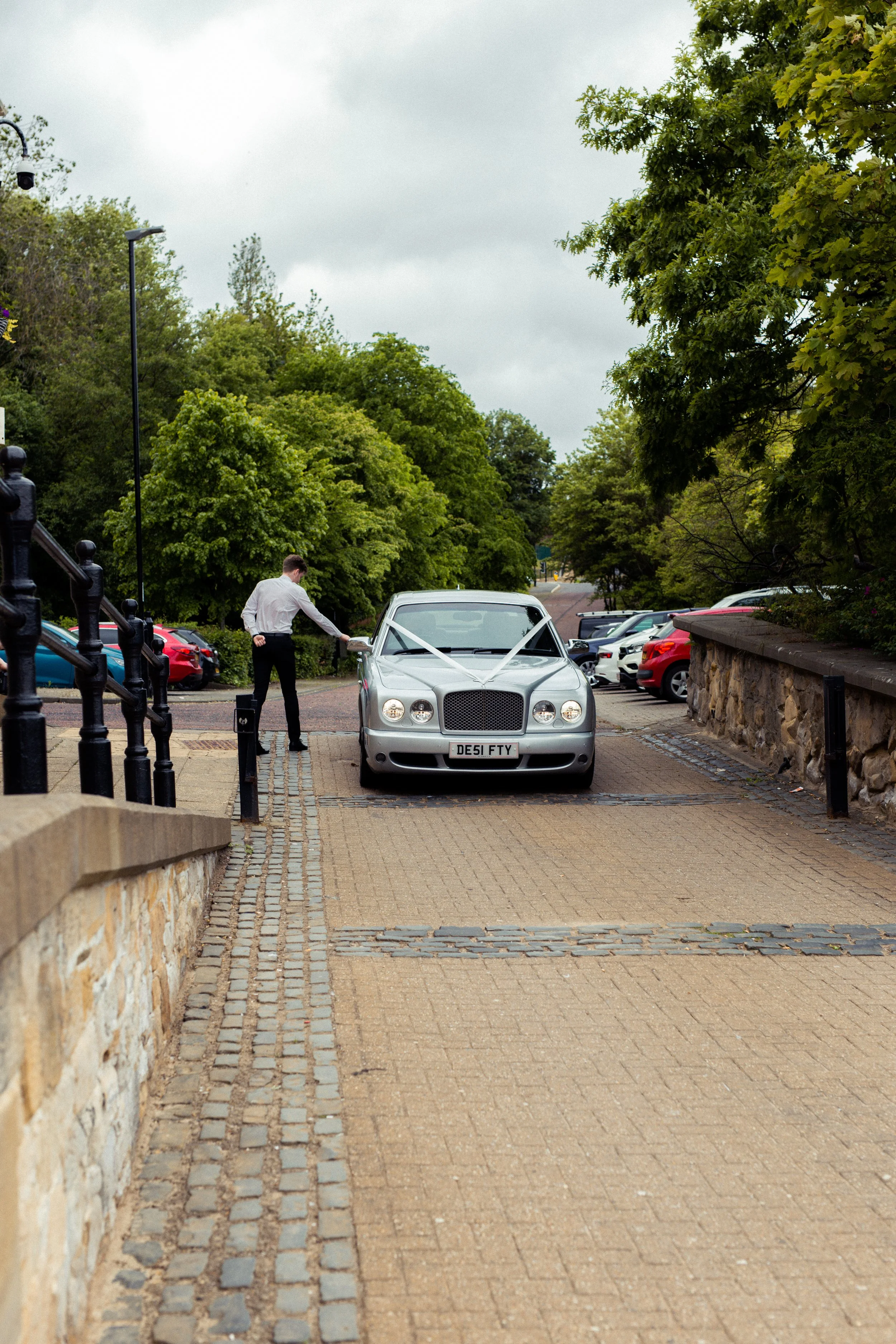 A man in formal attire is placing a ribbon with a bow on a silver luxury car, indicating a wedding or special occasion, on a paved pathway surrounded by trees and parked cars.