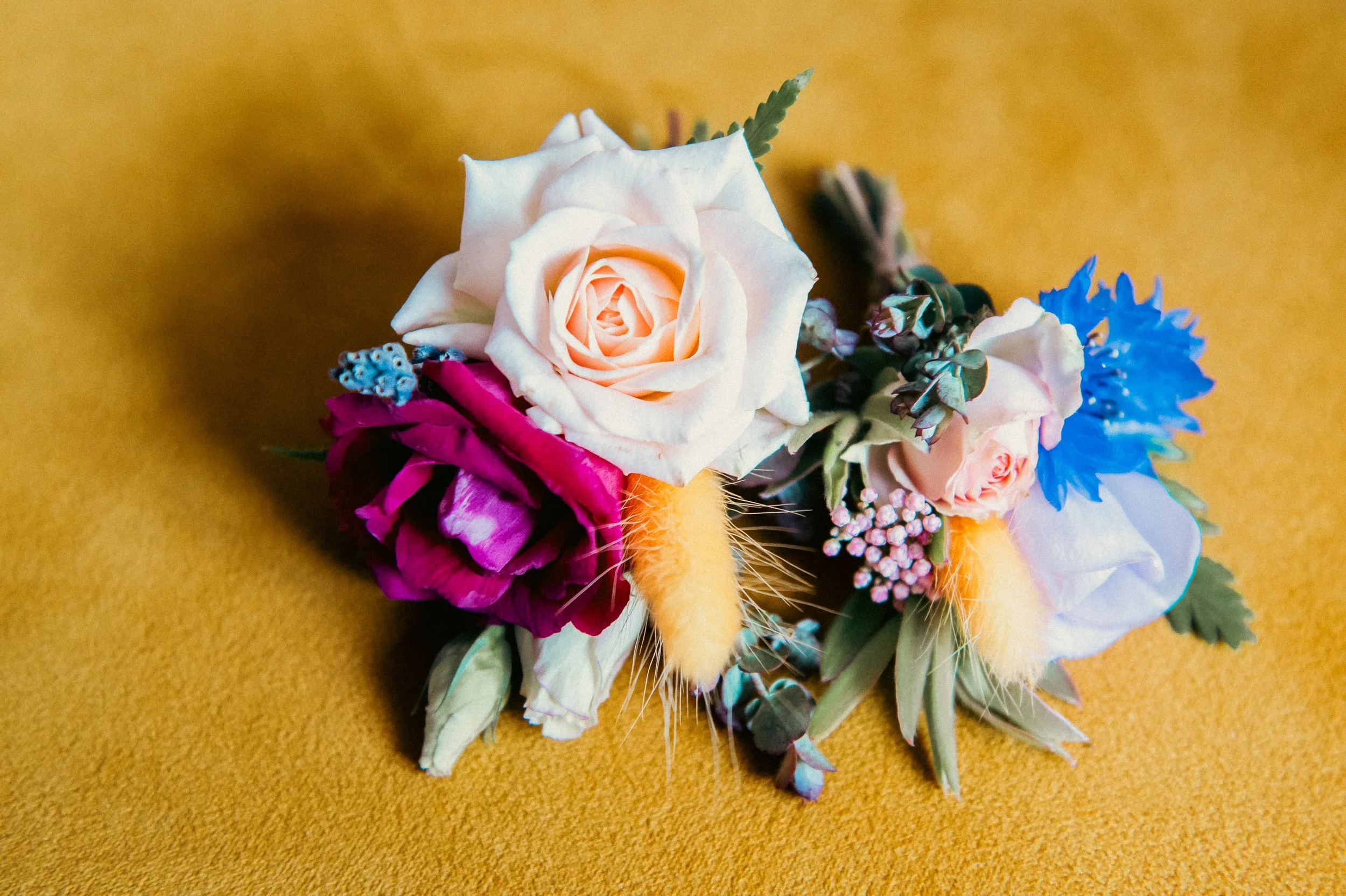 A floral arrangement featuring white roses, a dark pink rose, small blue and pink flowers, green leaves, and orange fuzzy accents on a mustard yellow background.