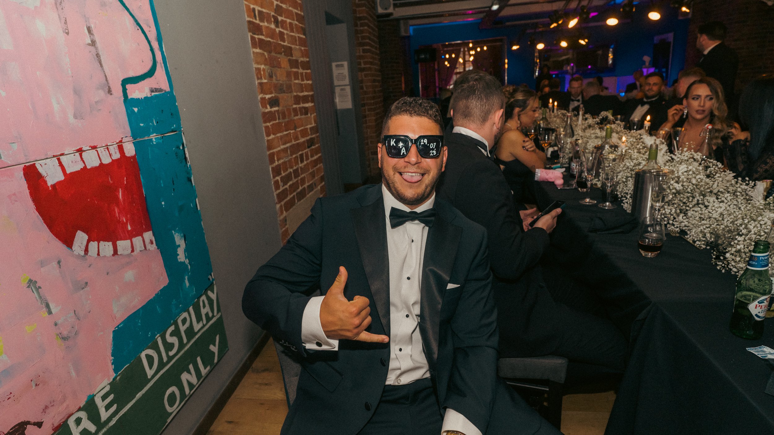 A man in a tuxedo with sunglasses is smiling and giving a thumbs up at a formal dinner event.