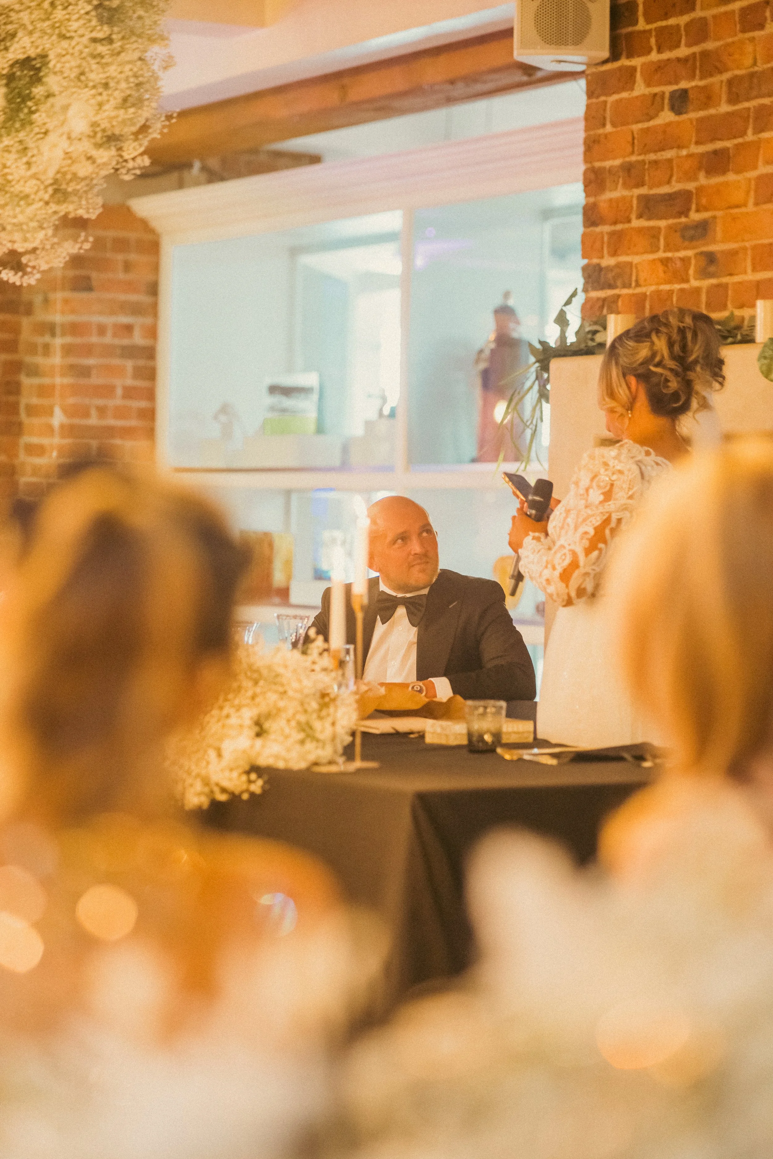 A man in a tuxedo looking at a woman giving a speech with a microphone at a wedding reception.