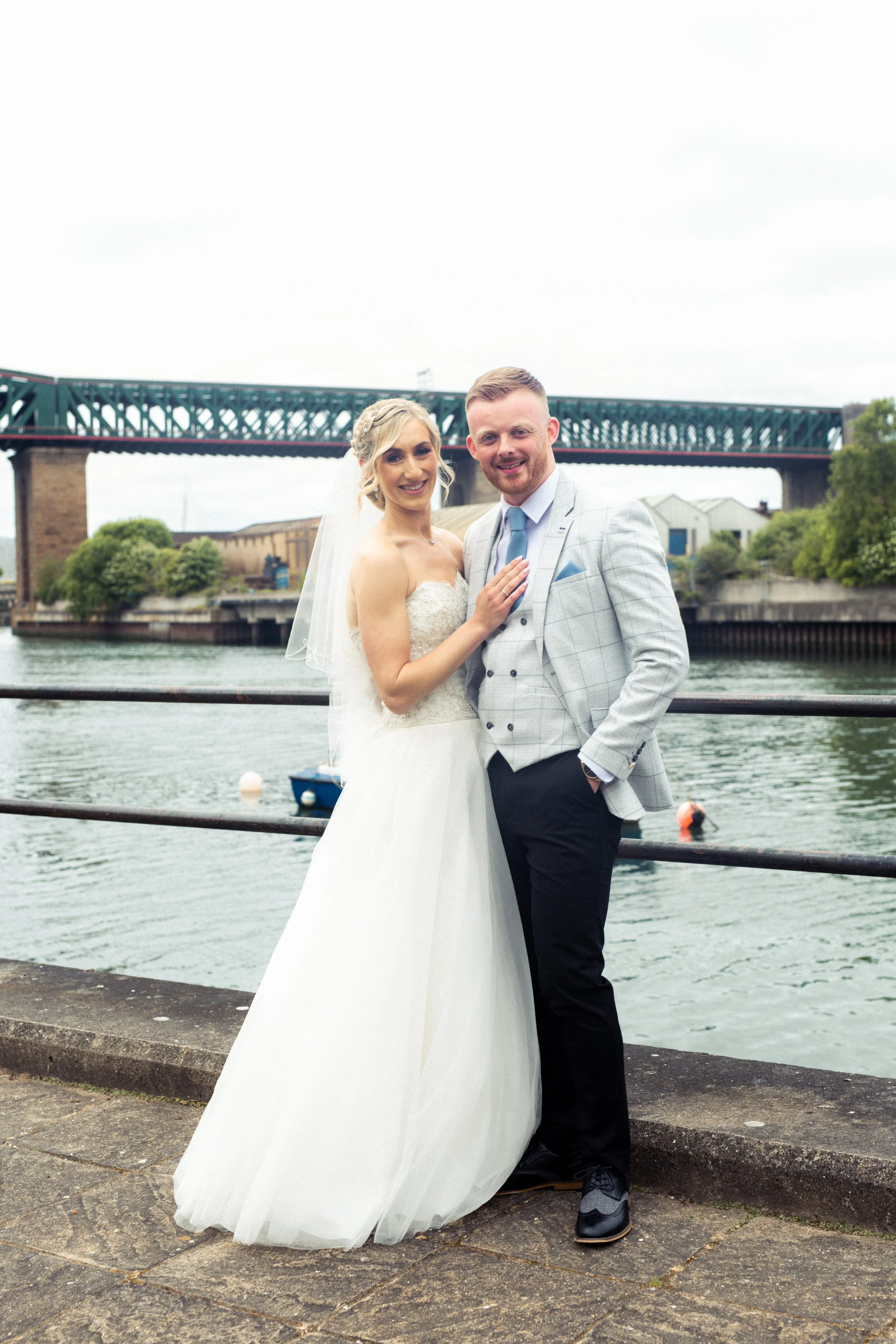 A bride and groom dressed in wedding attire standing by the water with a bridge in the background.