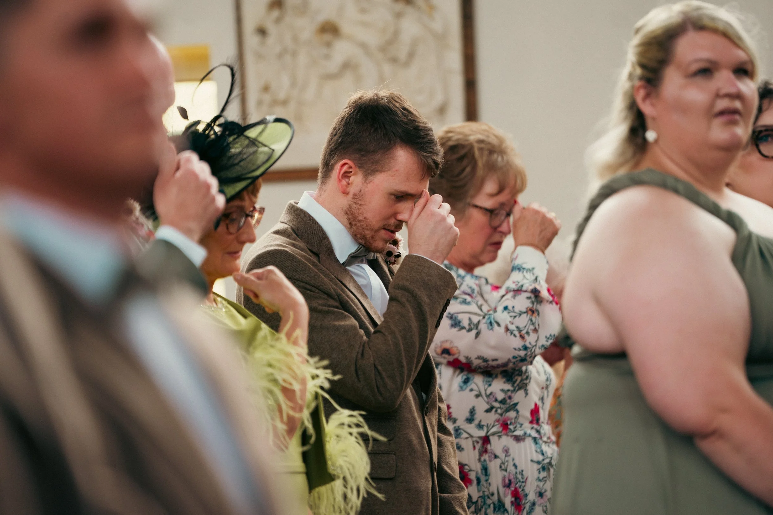 People at a formal gathering, some with eyes closed and hands clasped, appearing to pray or reflect in a solemn moment.