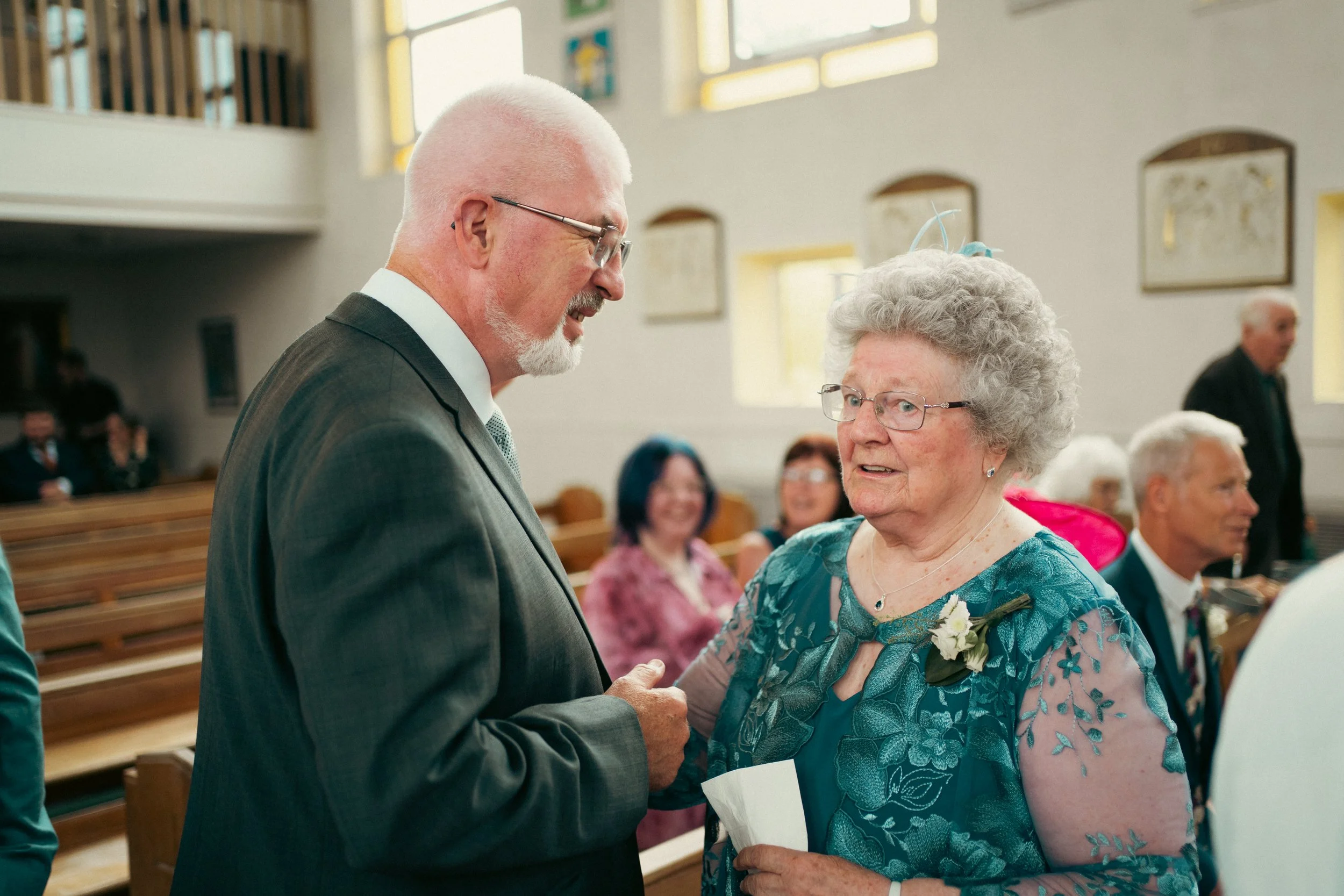 An elderly woman with gray, curly hair and glasses, wearing a teal lace dress with a white floral boutonniere, engaging in conversation with a man in a gray suit inside a church during a wedding.