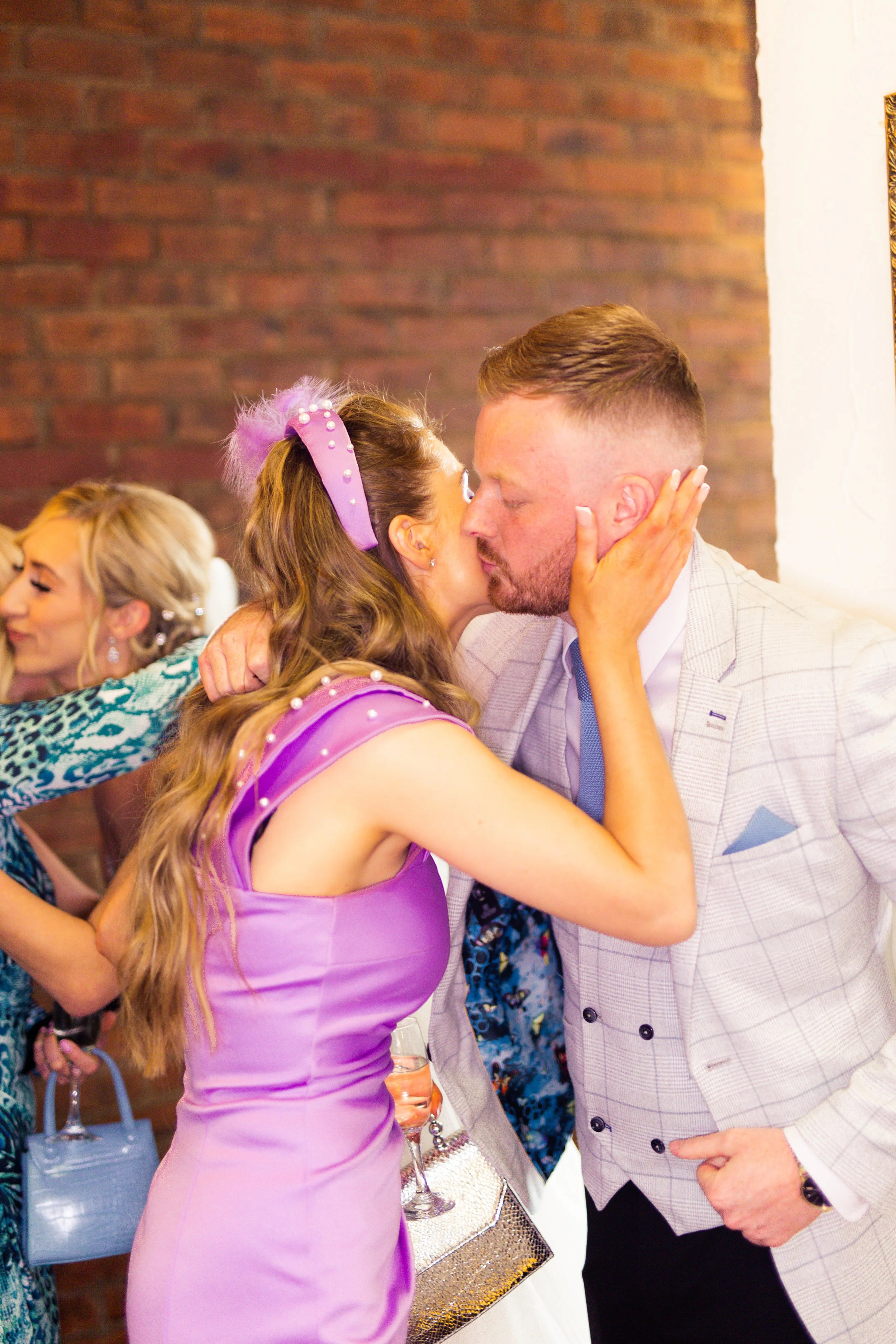 A woman in a purple dress and a man in a gray suit sharing a kiss at a celebration, with other women in colorful dresses in the background.