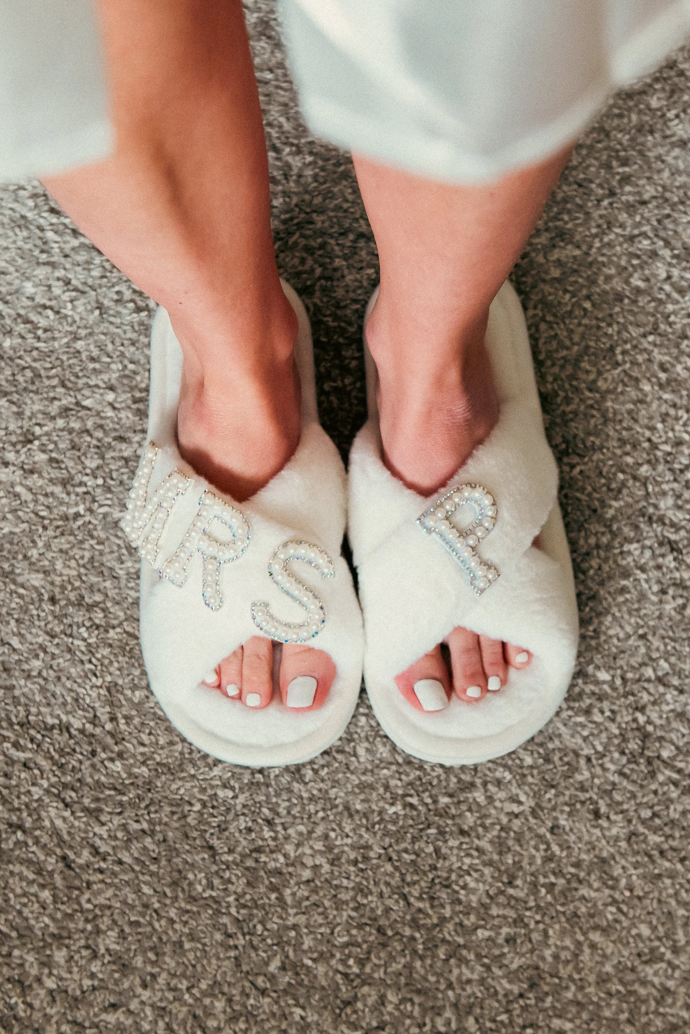 A person standing on a carpeted floor wearing fluffy white slippers with the words 'HURTS' and 'R' spelled out in rhinestones on each slipper, and painted white toenails.