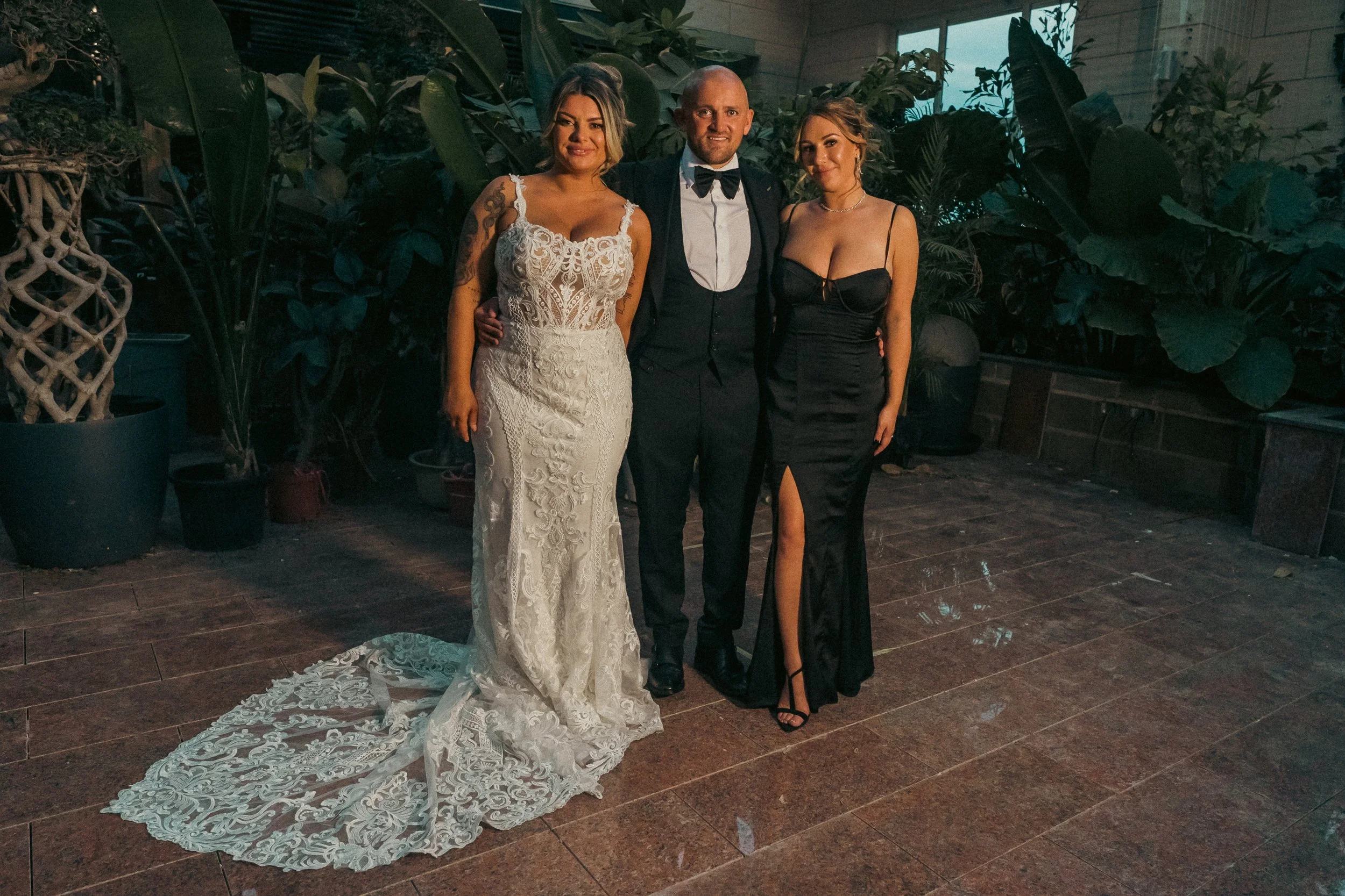 Three people standing together, two women in elegant black dresses and a man in a tuxedo, in a setting with large green plants and a tiled floor.
