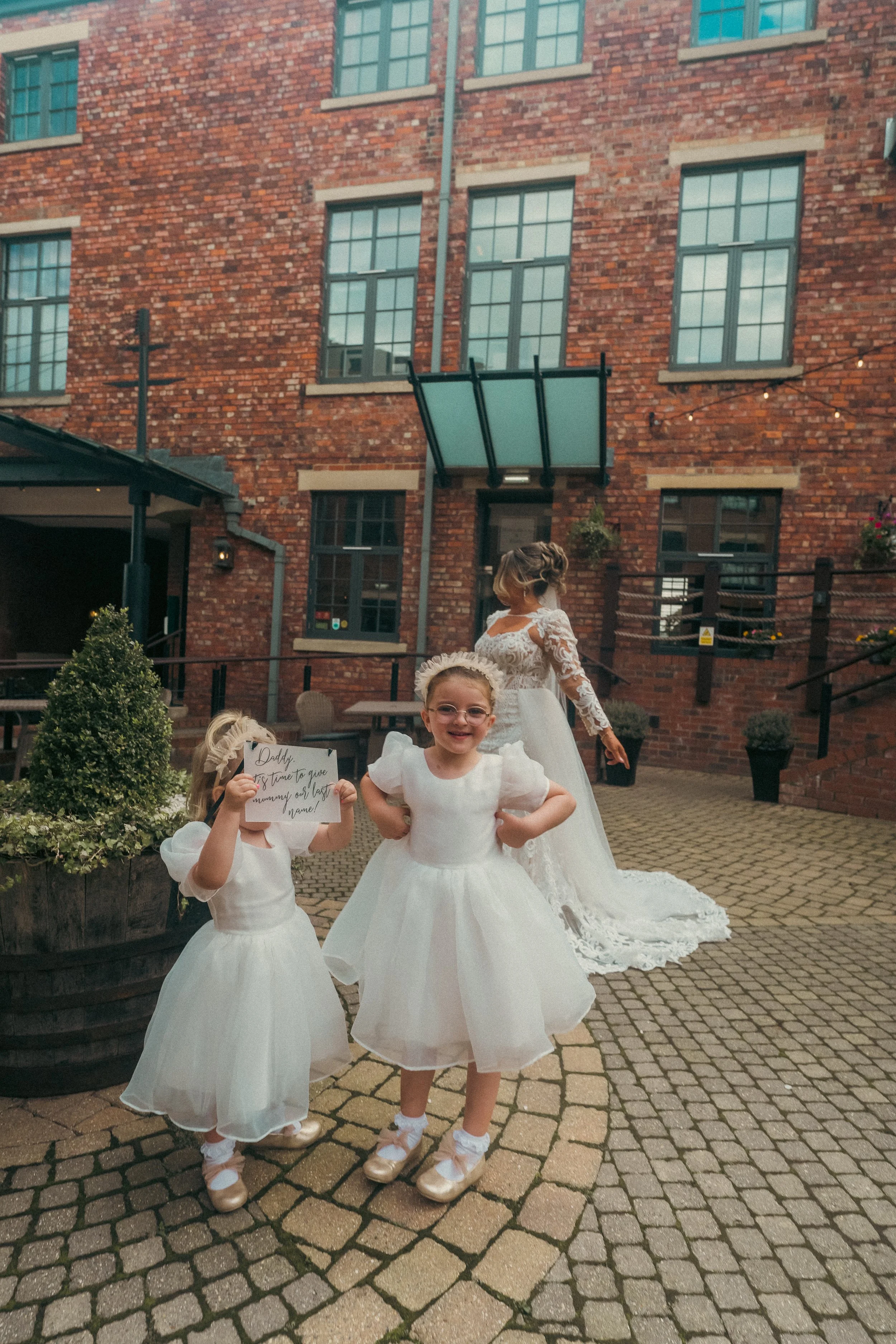 A bride in a wedding dress stands in the background in an outdoor courtyard, with two young girls in white dresses and gold shoes in the foreground. One girl is holding a sign that reads 'Daddy, it's time to give mummy our last name!' and all are smi