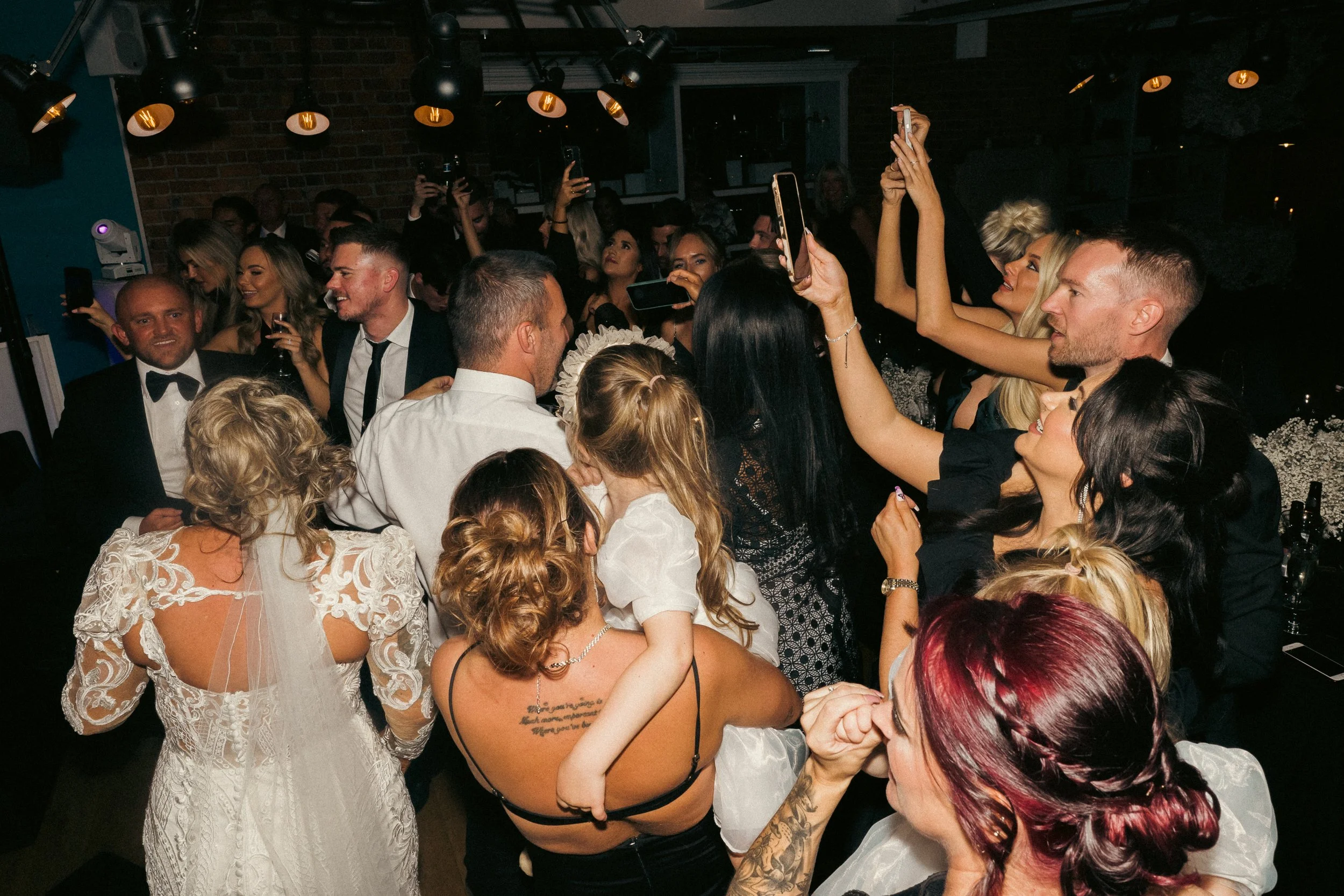 A large group of people at a party or celebration, some taking photos or videos, mingling and smiling, in a dimly lit venue with brick walls and hanging lights.