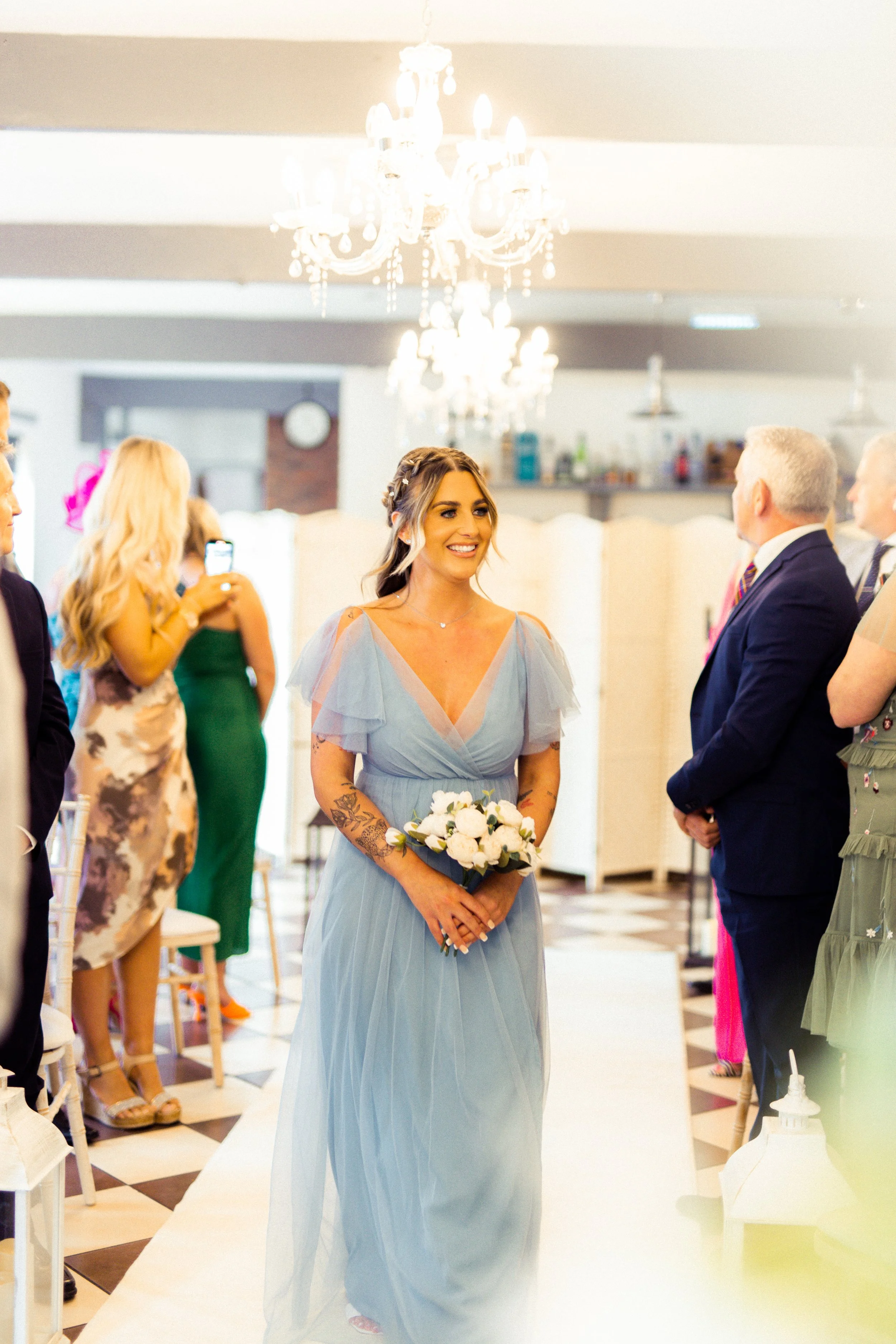 A bride in a light blue dress holding a bouquet walks down the aisle at her wedding ceremony, surrounded by guests in a decorated indoor venue with chandeliers.