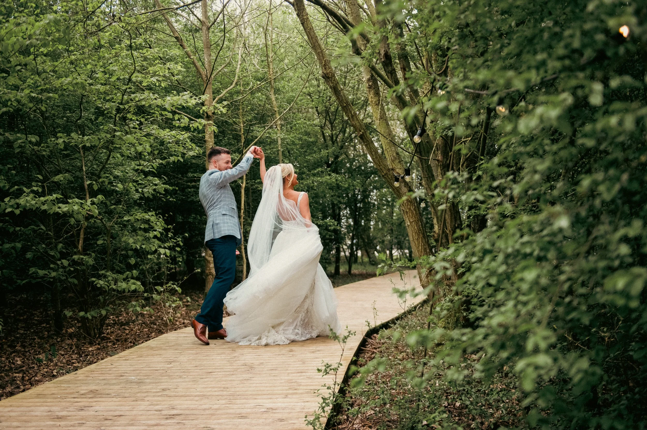 A newlywed couple is dancing on a wooden pathway in a lush green forest, with string lights hanging from the trees.