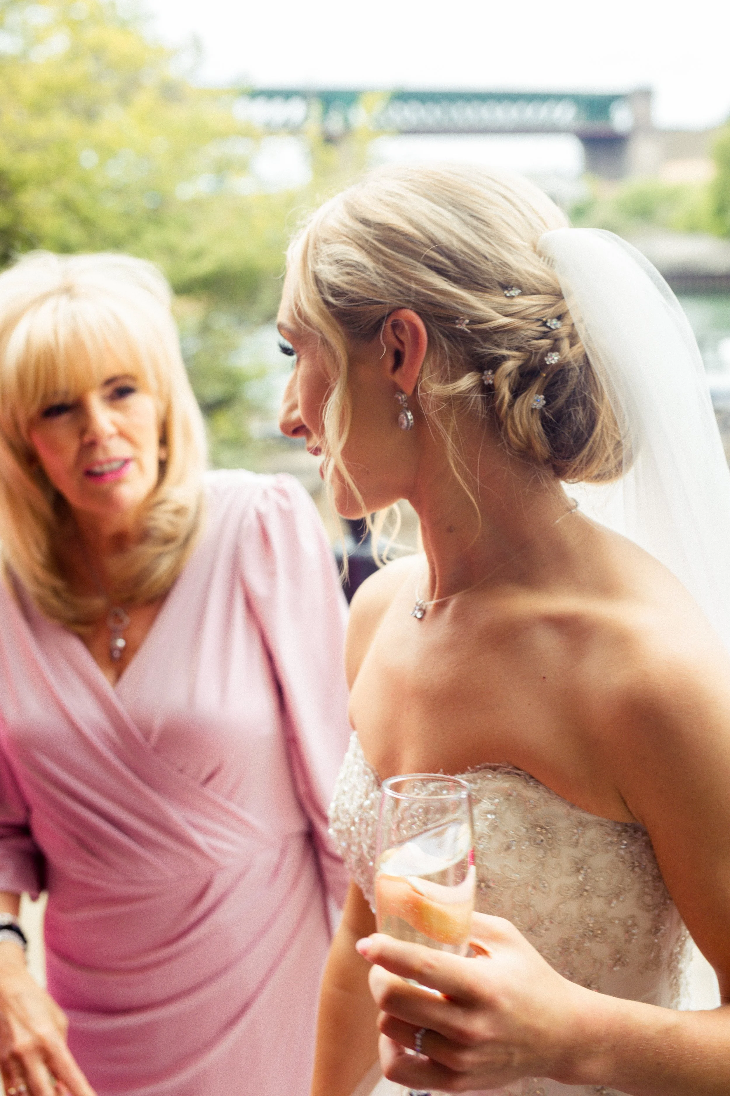 A bride with an elaborate hairstyle and earrings holding a glass of champagne, smiling and engaging in conversation with an older woman wearing a pink dress with blonde hair.
