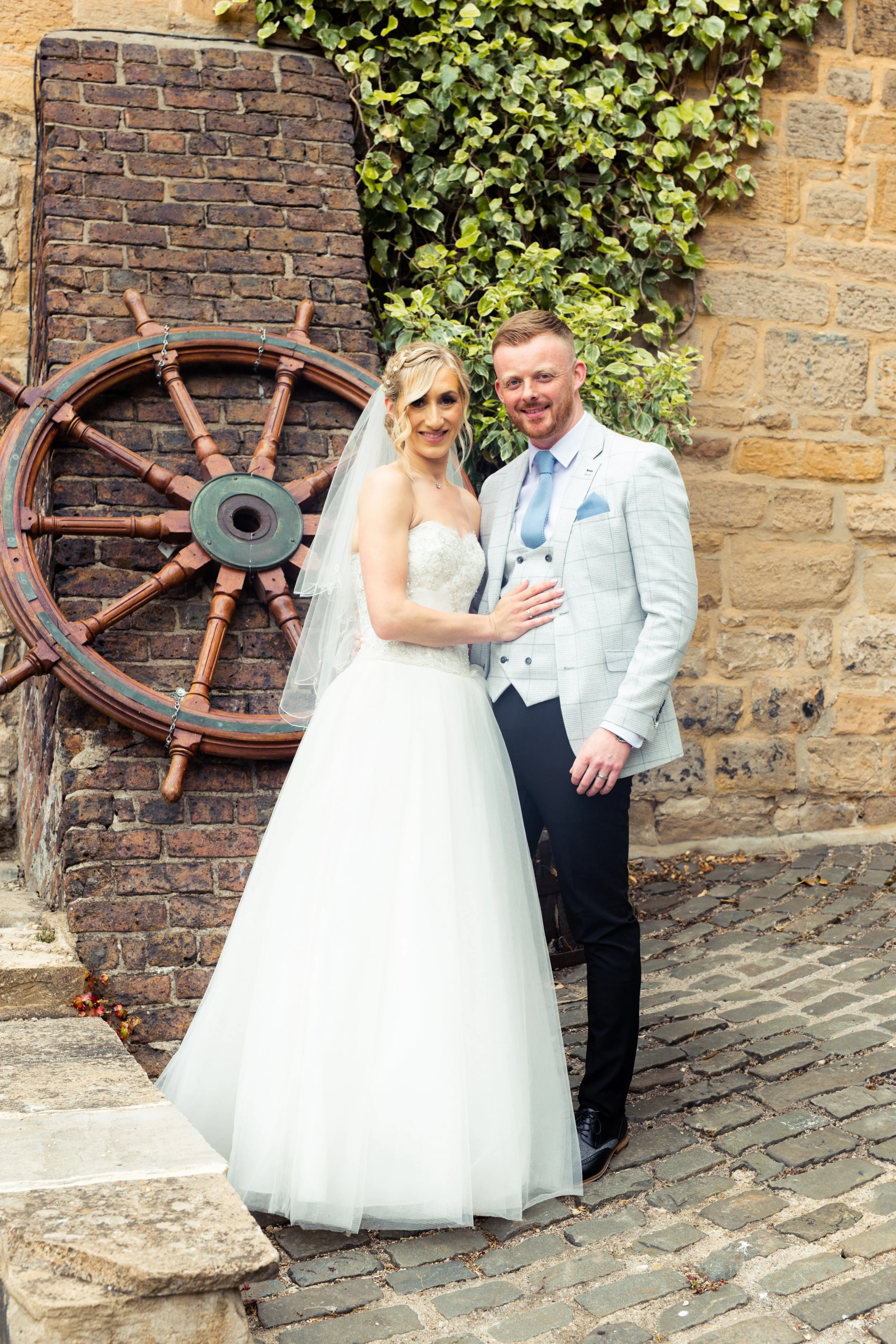 A bride and groom posing outdoors near a brick wall with an old ship's steering wheel and green ivy.