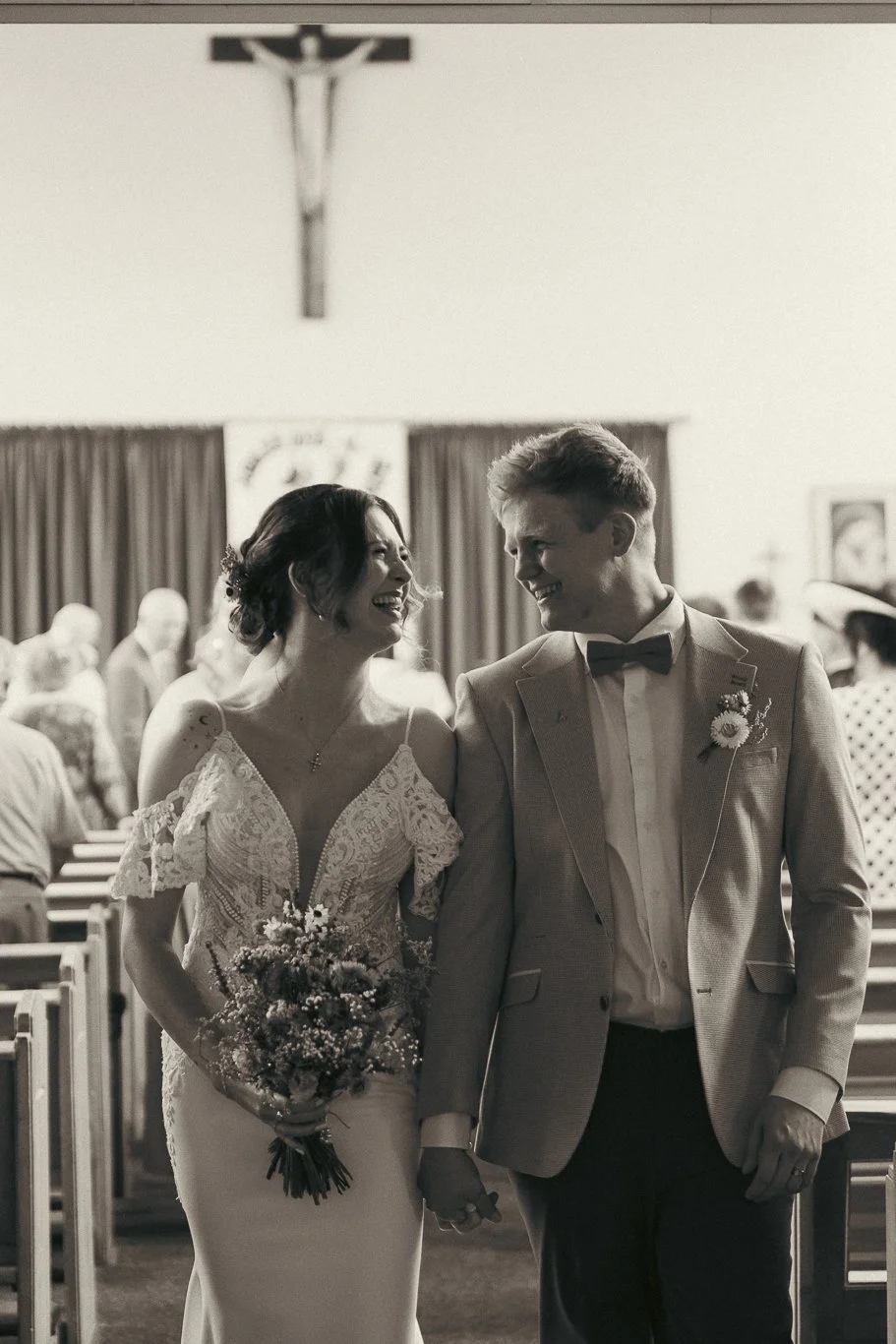 Black and white photo of a bride and groom smiling at each other during their wedding ceremony inside a church, with guests seated in pews in the background.