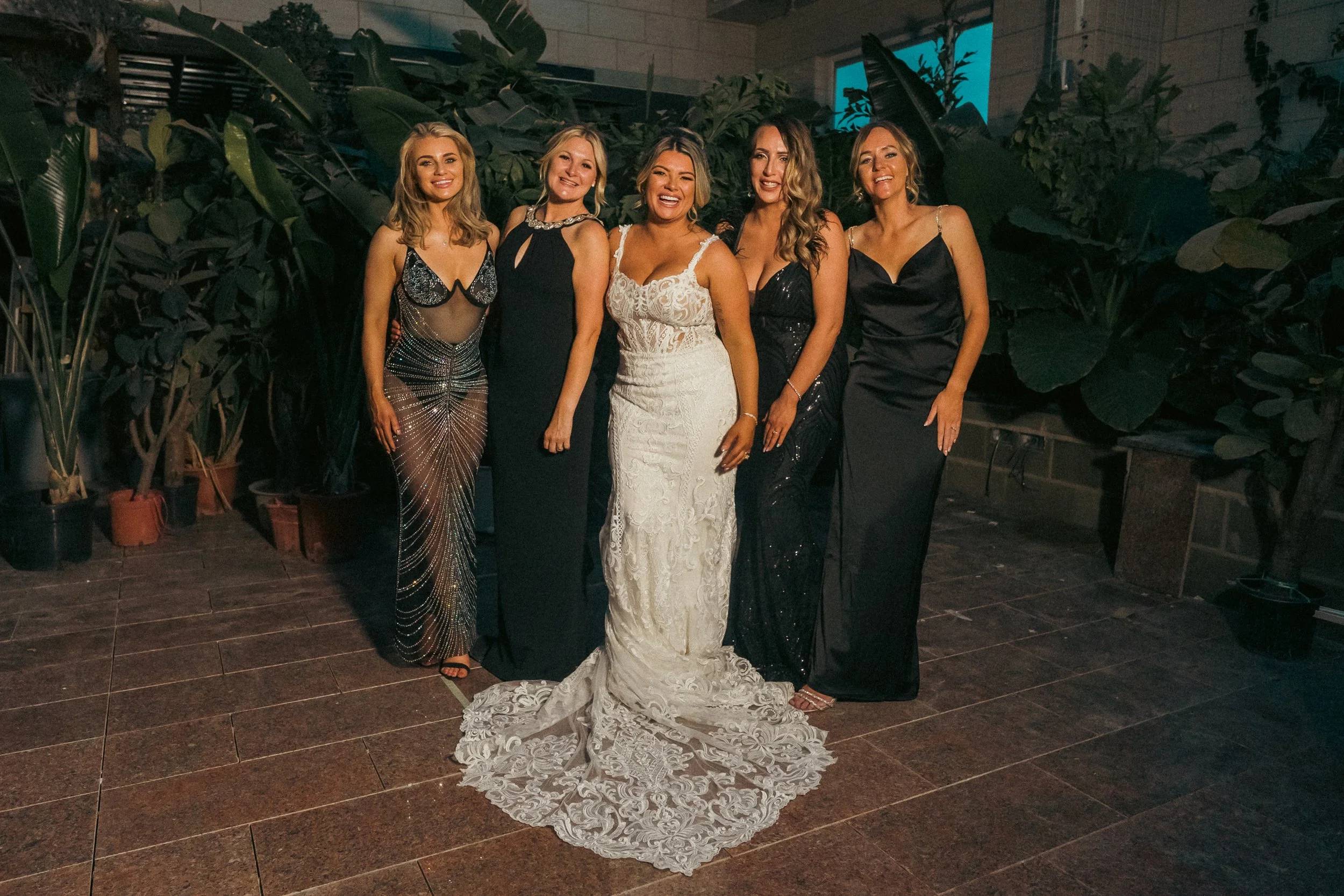 Group of six women in formal dresses posing indoors with plants in background.