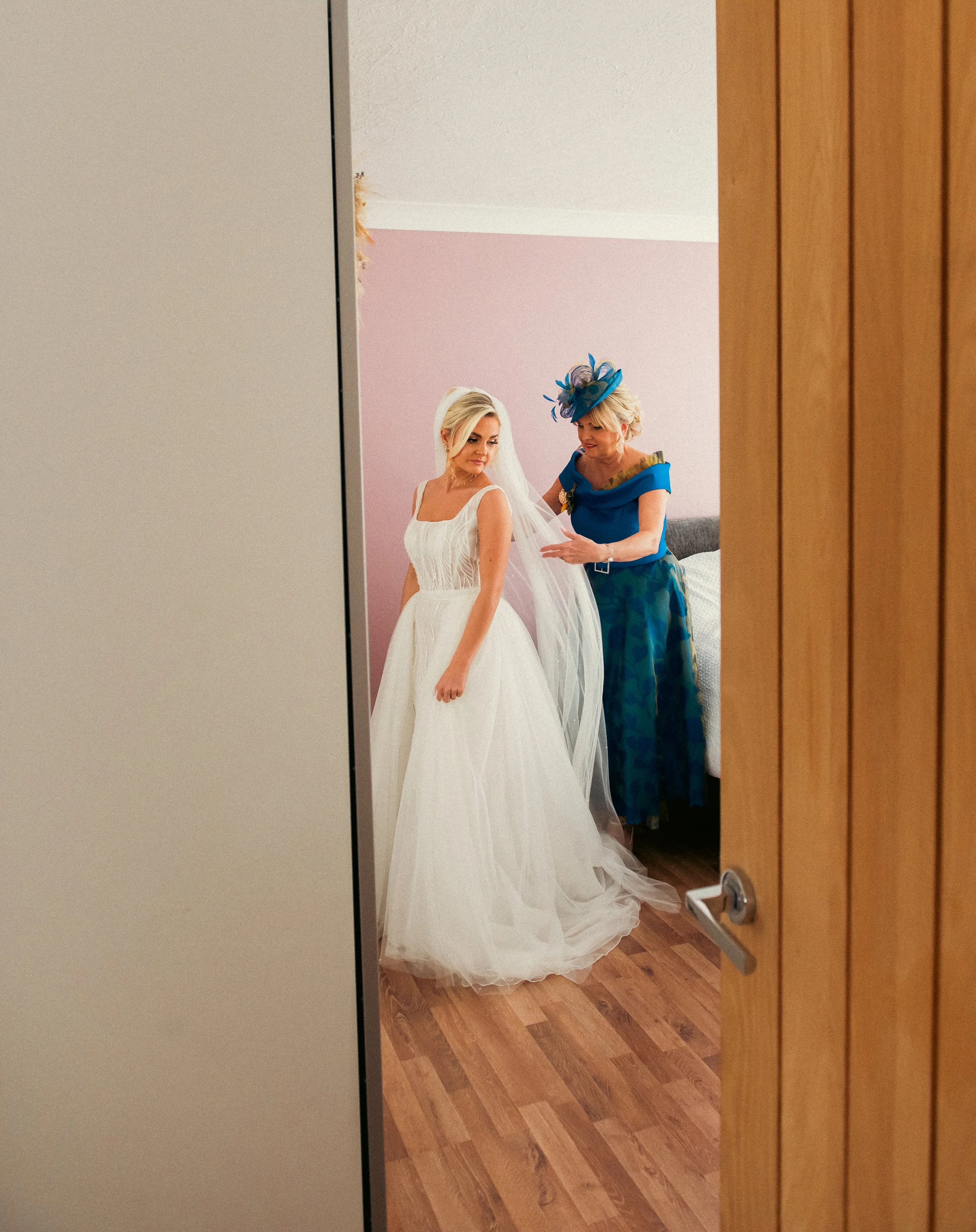 A bride in a white wedding gown stands while an older woman, possibly her mother, helps her with her veil in a bedroom.