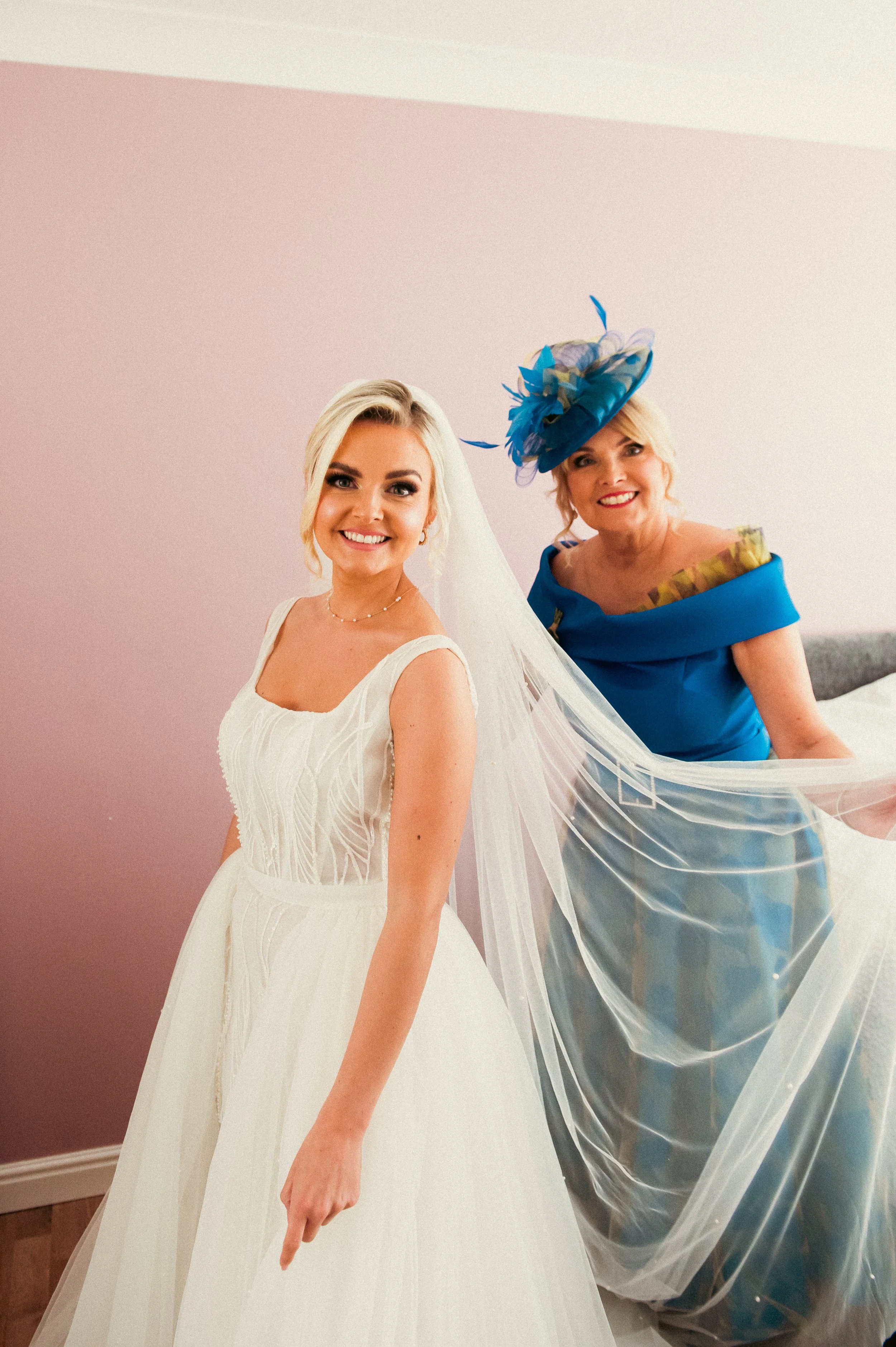 A bride in a white wedding dress and veil smiling at the camera, standing beside an older woman in a blue dress and elaborate hat, holding a sheer fabric.