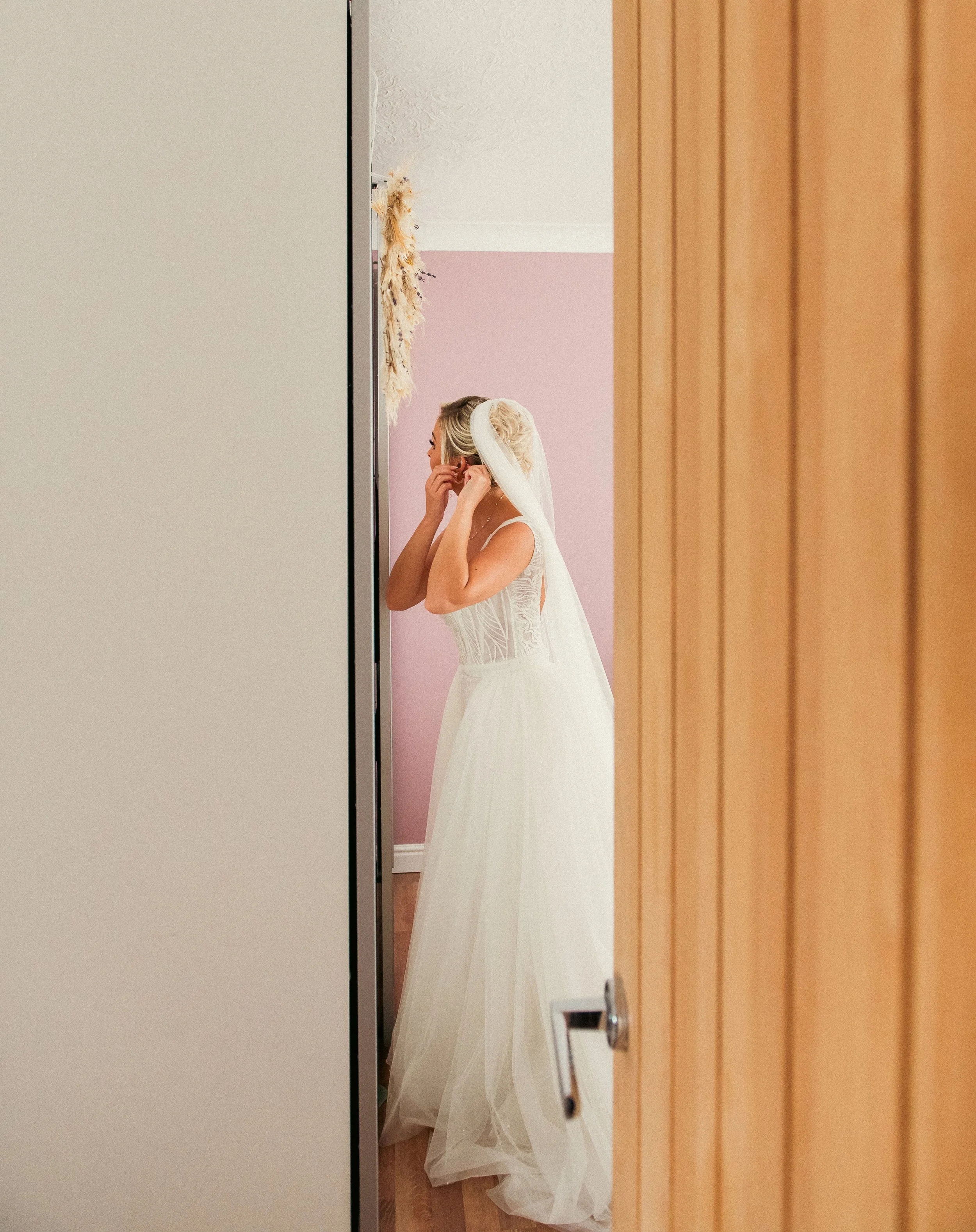 Bride in a wedding dress adjusting her earrings in a mirror, seen through partly open closet doors.