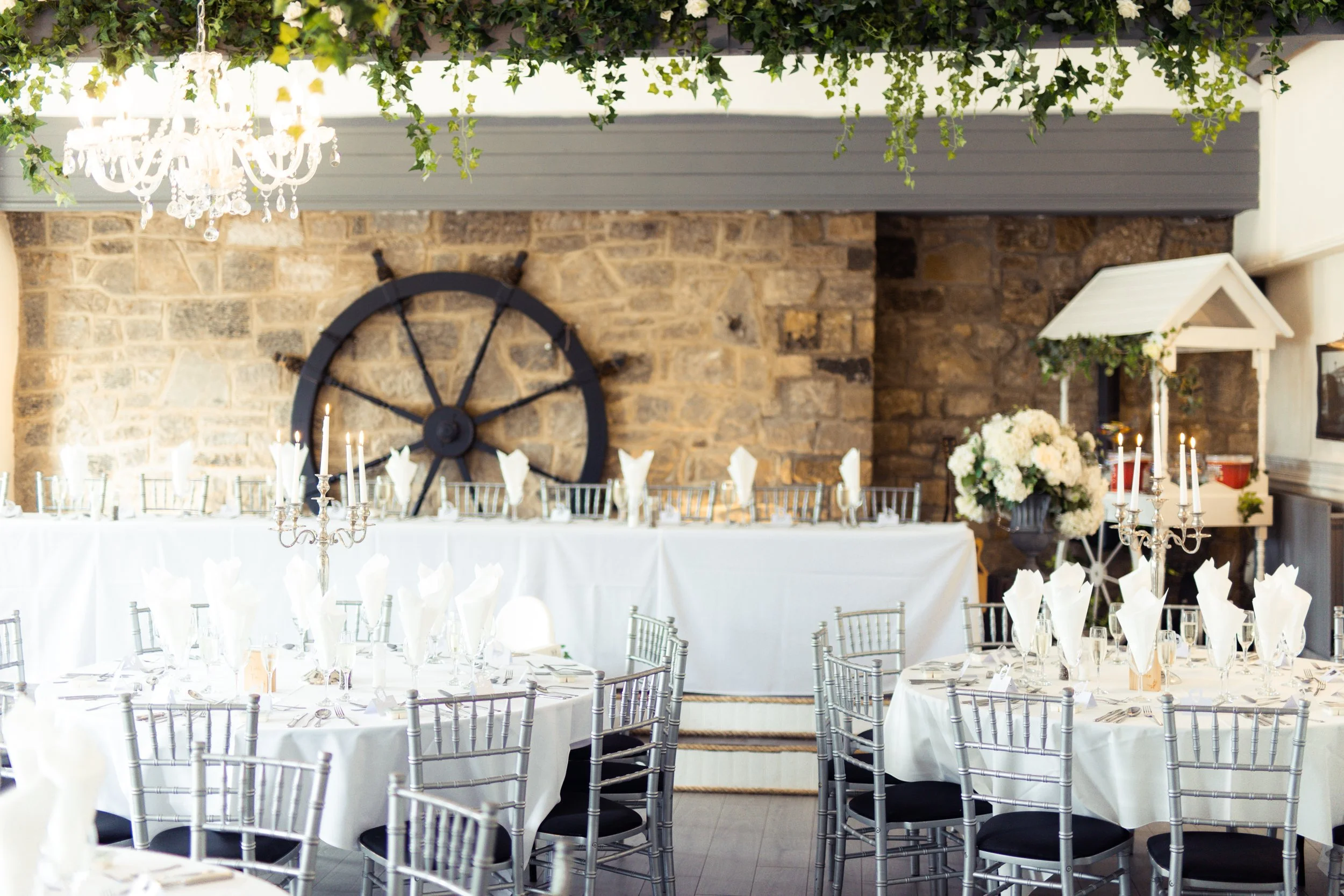 Elegant banquet hall with white tablecloths, silver chairs, tall candelabras with lit candles, and a floral centerpiece. A clock with candles is mounted on a brick wall under a hanging chandelier with greenery overhead.