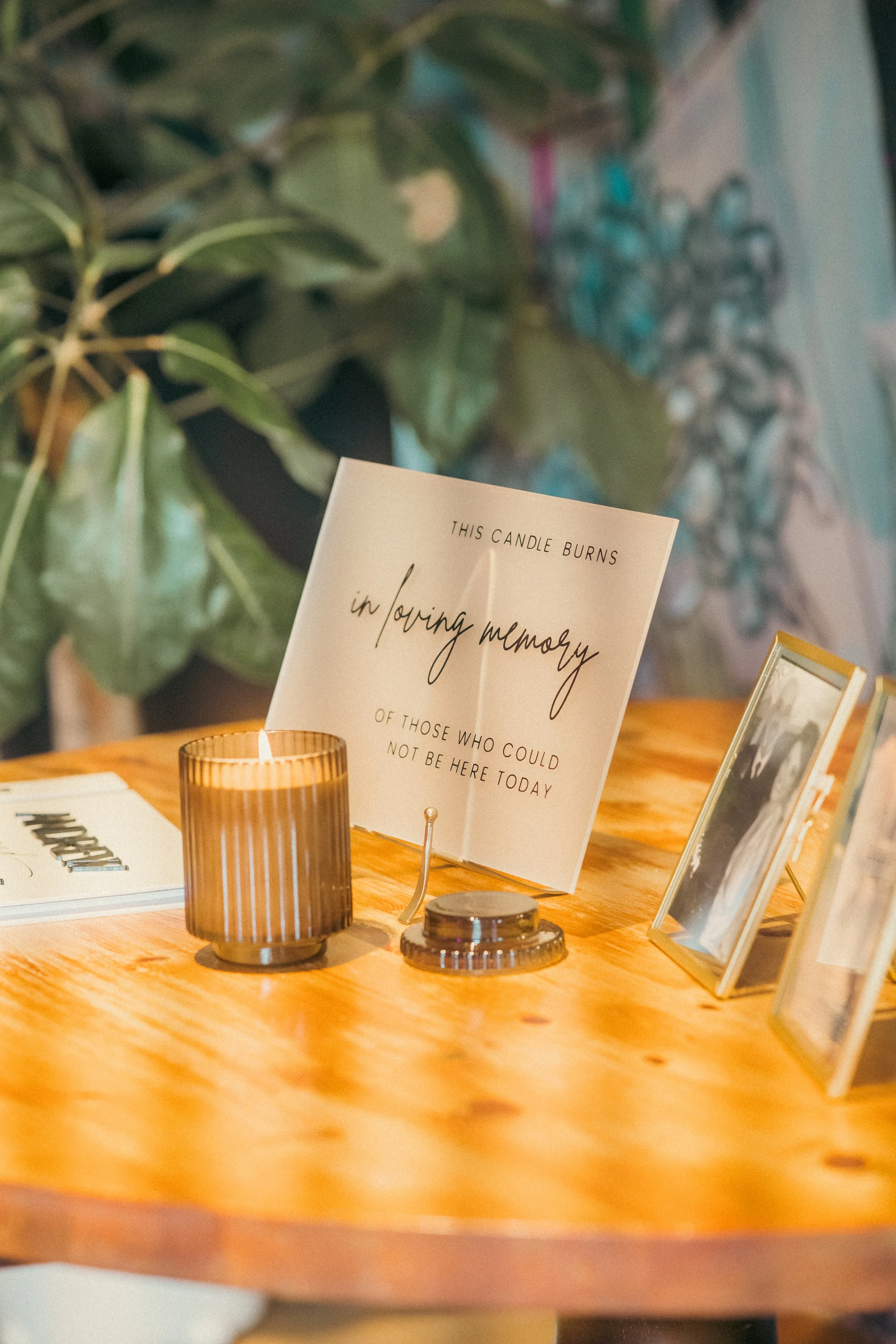 A memorial display on a wooden table with a candle, framed photographs, and a sign that reads 'This candle burns in loving memory of those who could not be here today.'