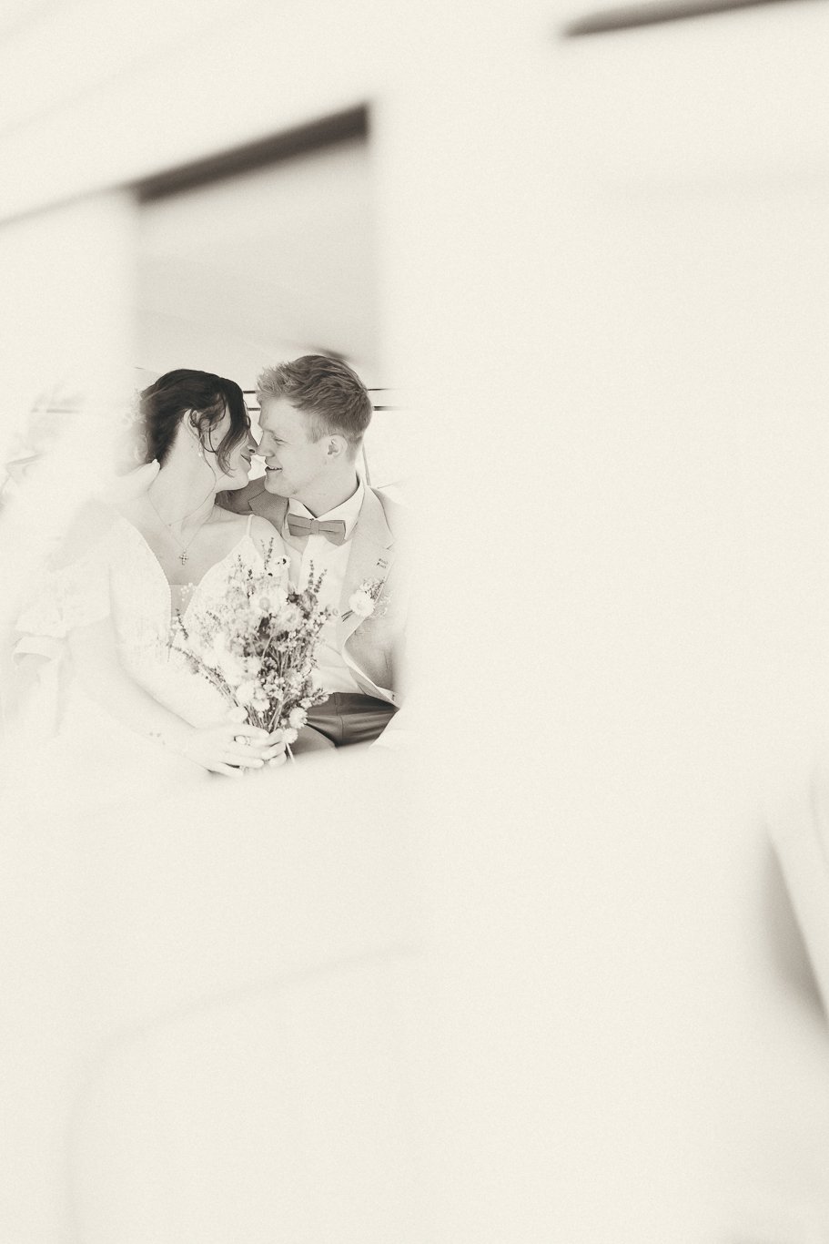 Black and white photo of a couple on their wedding day, smiling at each other very closely, holding a bouquet of flowers.