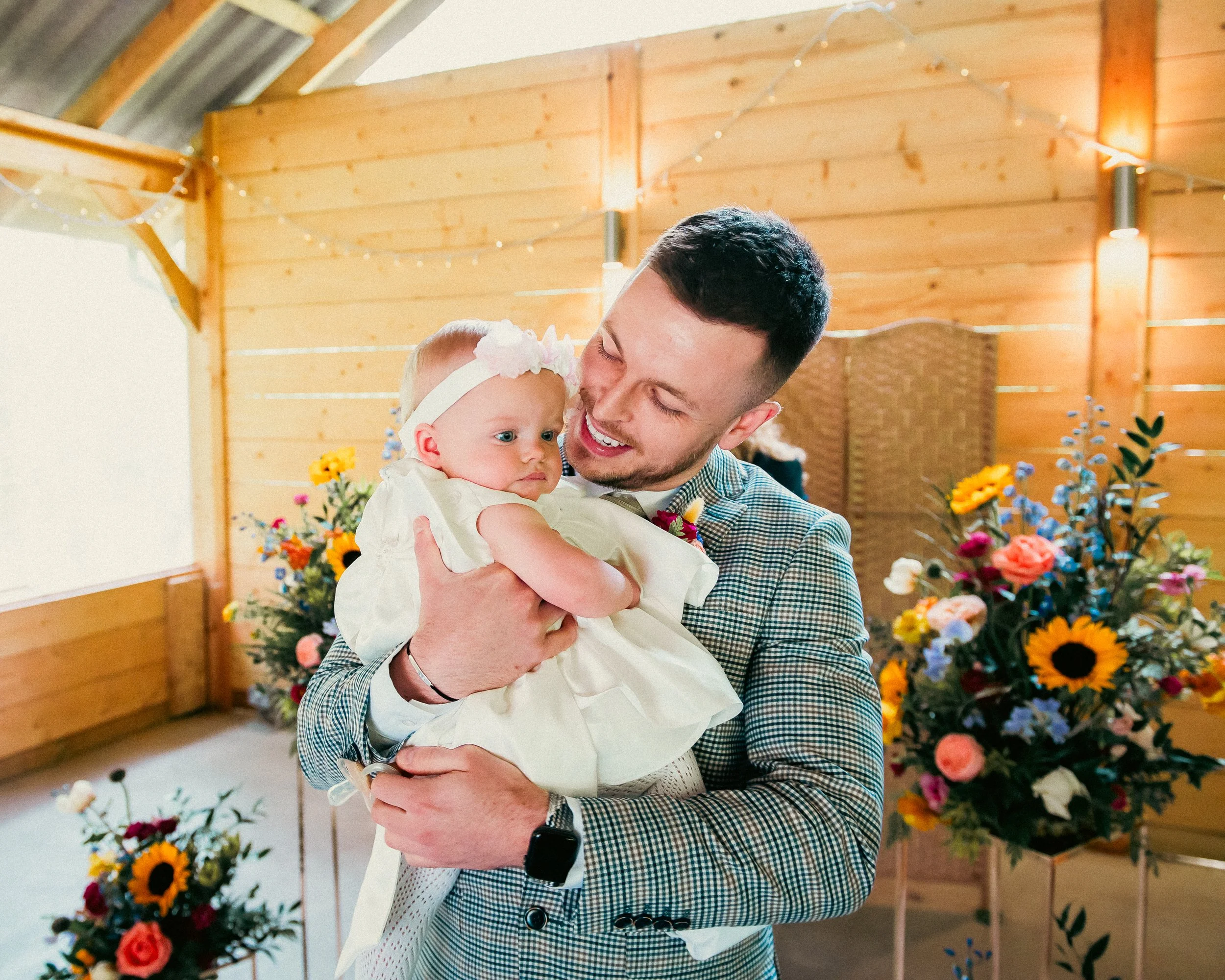 A man holding a baby girl in a decorated indoor setting with flowers and string lights.