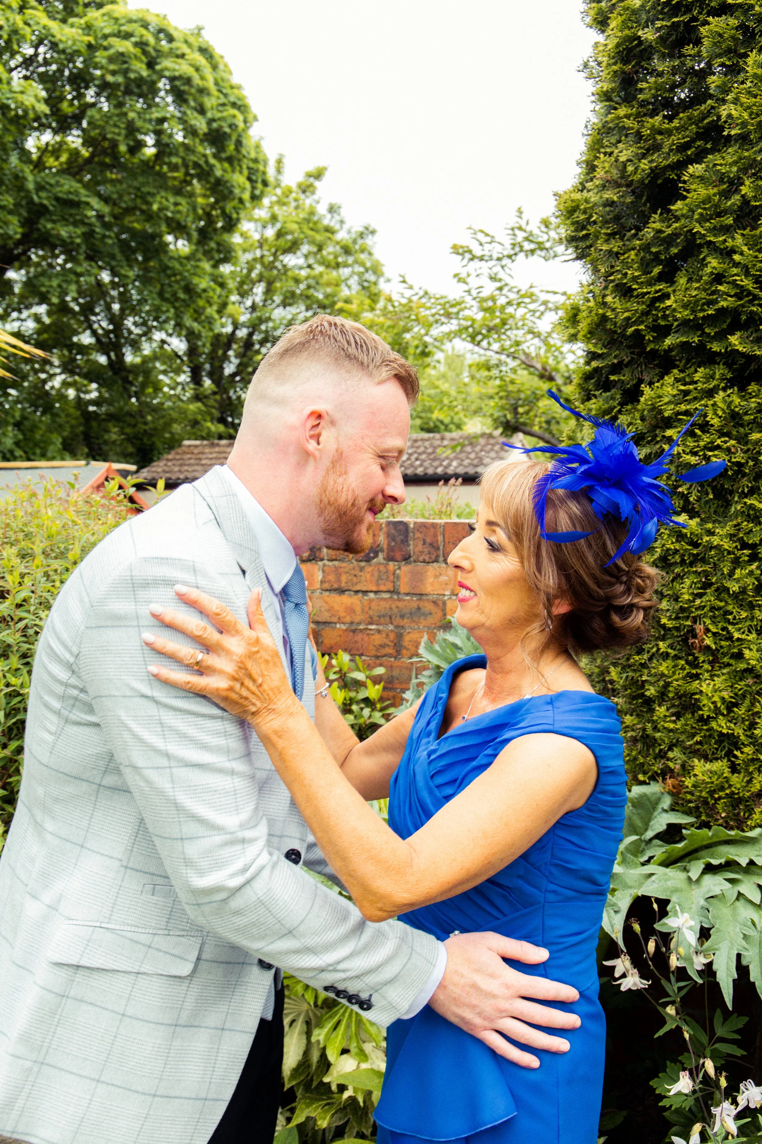 A young man and an older woman in blue dress and fascinator share a joyful moment outdoors, with greenery and a brick wall in the background.