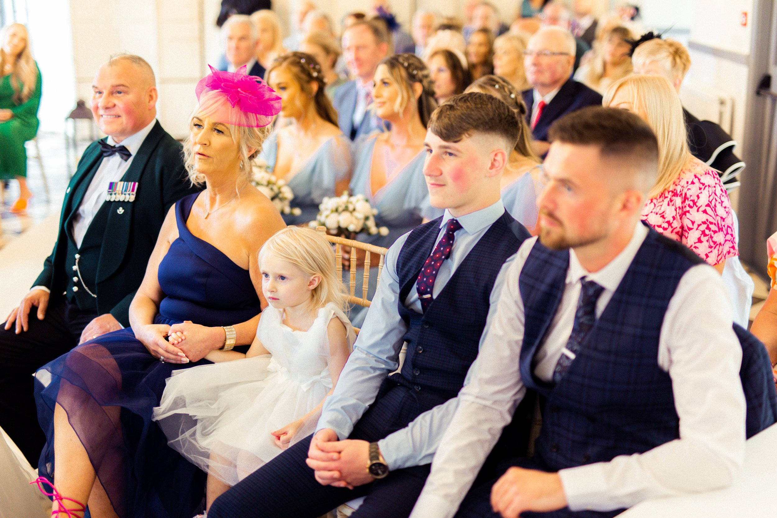People attending a wedding ceremony, seated in rows, with women in dresses and men in suits, a young girl in a white dress sitting next to an older woman in a dark blue dress wearing a pink fascinator.