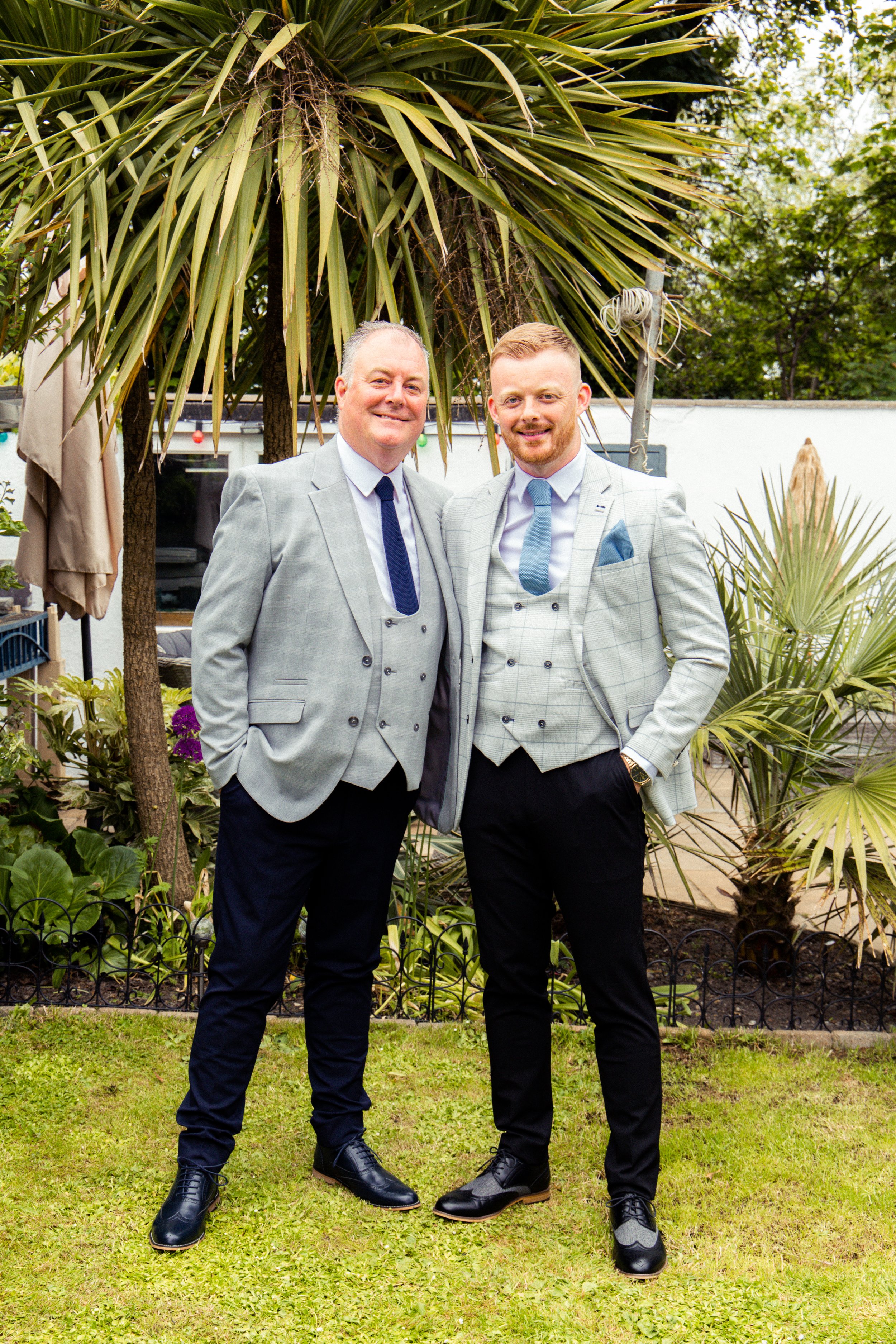 Two men in gray suits standing outdoors on grass, smiling, with trees and plants in the background.
