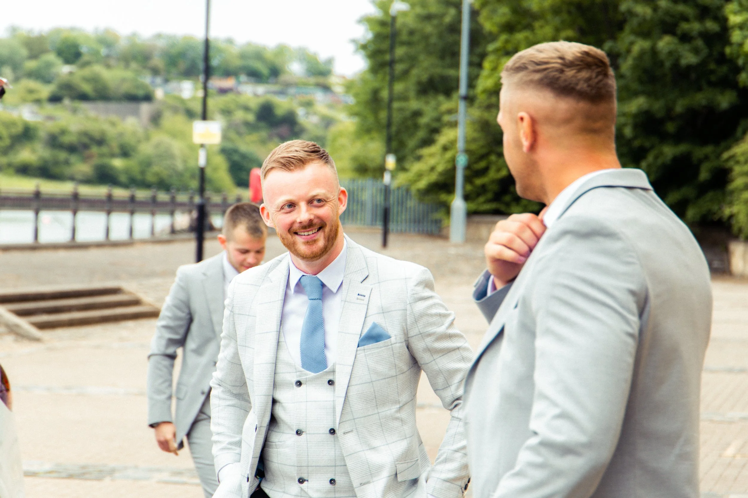 Three men dressed in gray suits talking outdoors near a river, with green trees and a pathway in the background.