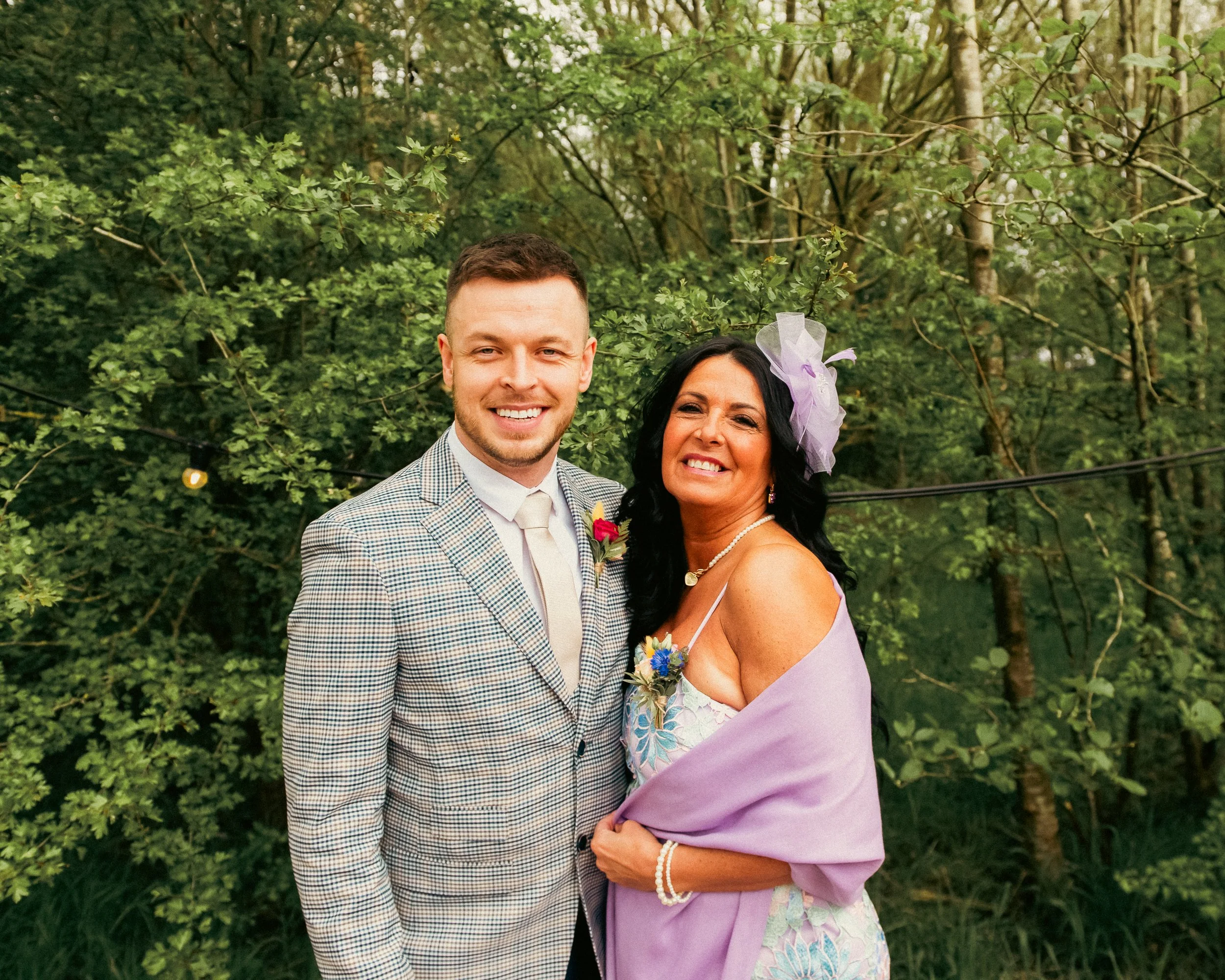 A young man and woman smiling outdoors, dressed in formal attire, surrounded by green trees and foliage.