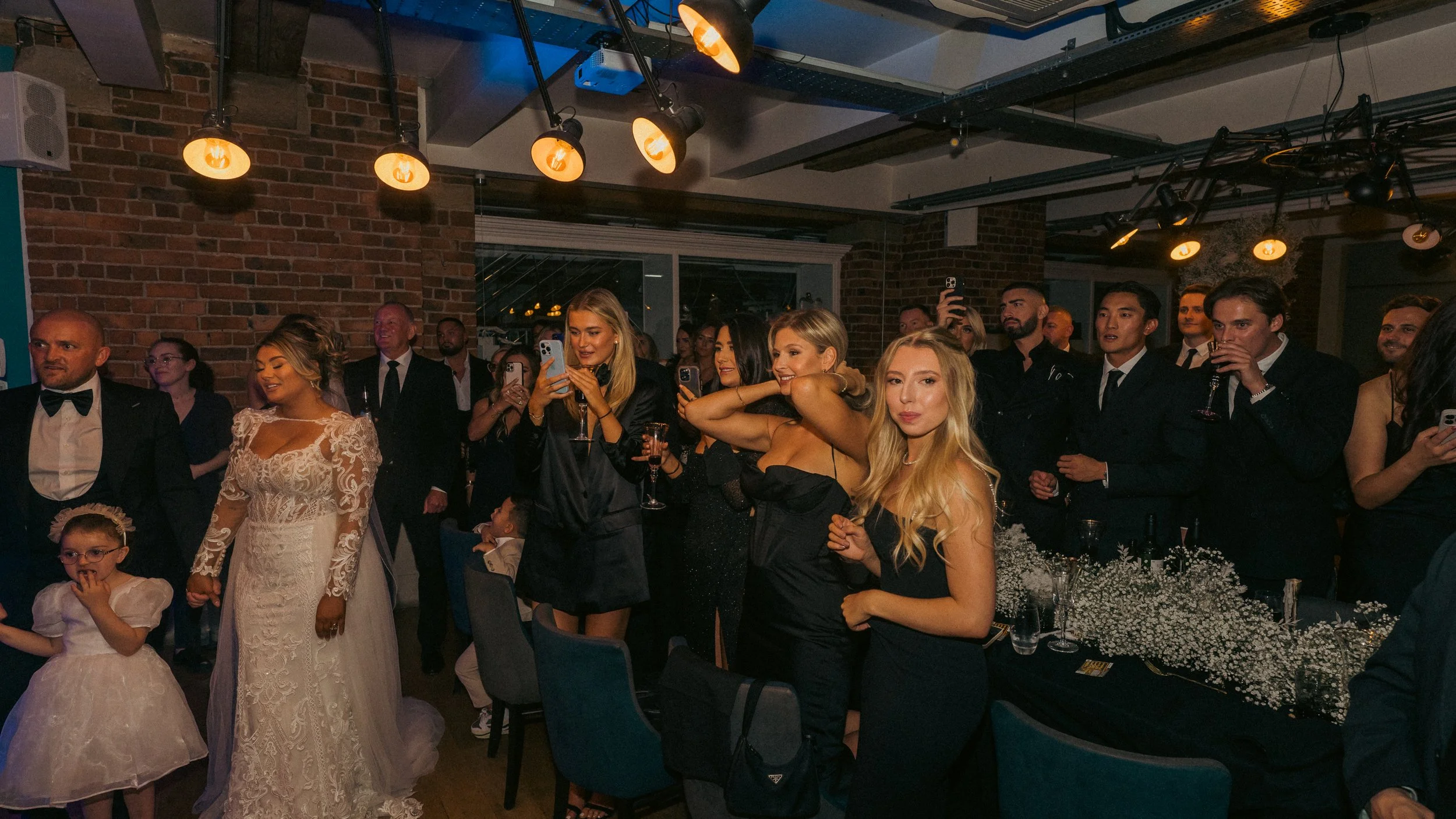 Group of people dressed in formal attire at an indoor event, some smiling, taking photos, and standing near tables decorated with flowers.