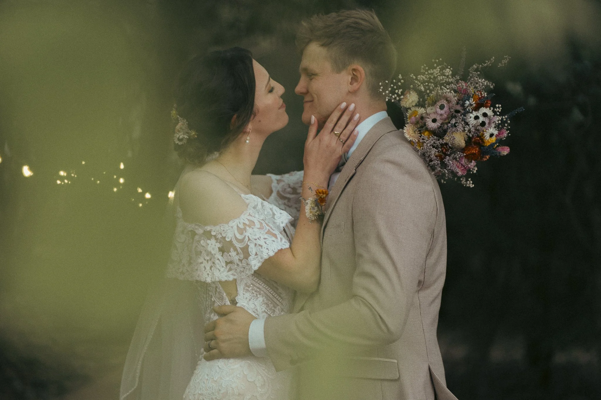 A bride and groom embrace and look into each other's eyes during their wedding, with the bride holding the groom's face and the groom holding the bride's waist, surrounded by trees and flowers.