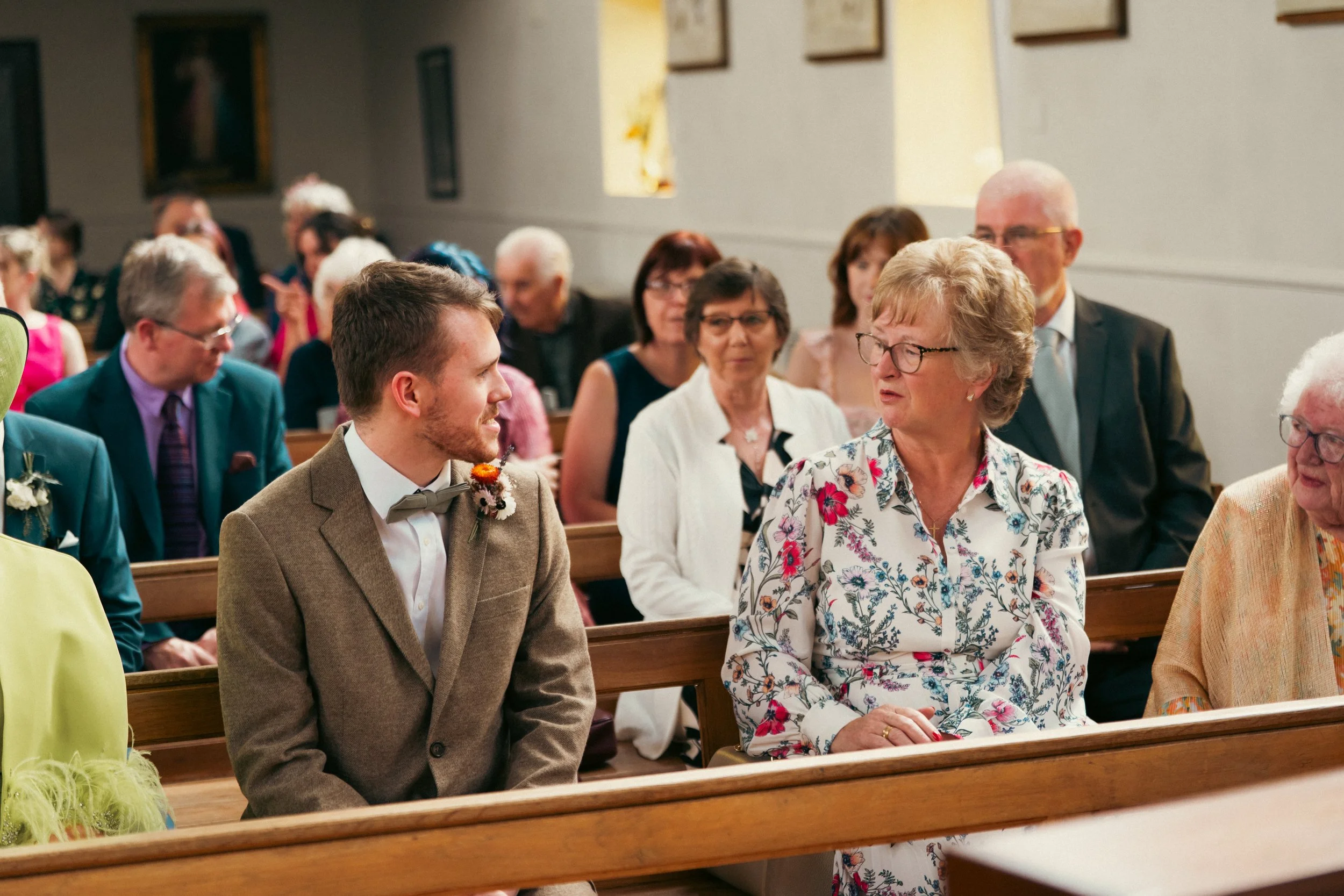 A wedding ceremony with guests seated in church pews, including a young man in a brown suit and bow tie talking to an older woman in a floral dress.