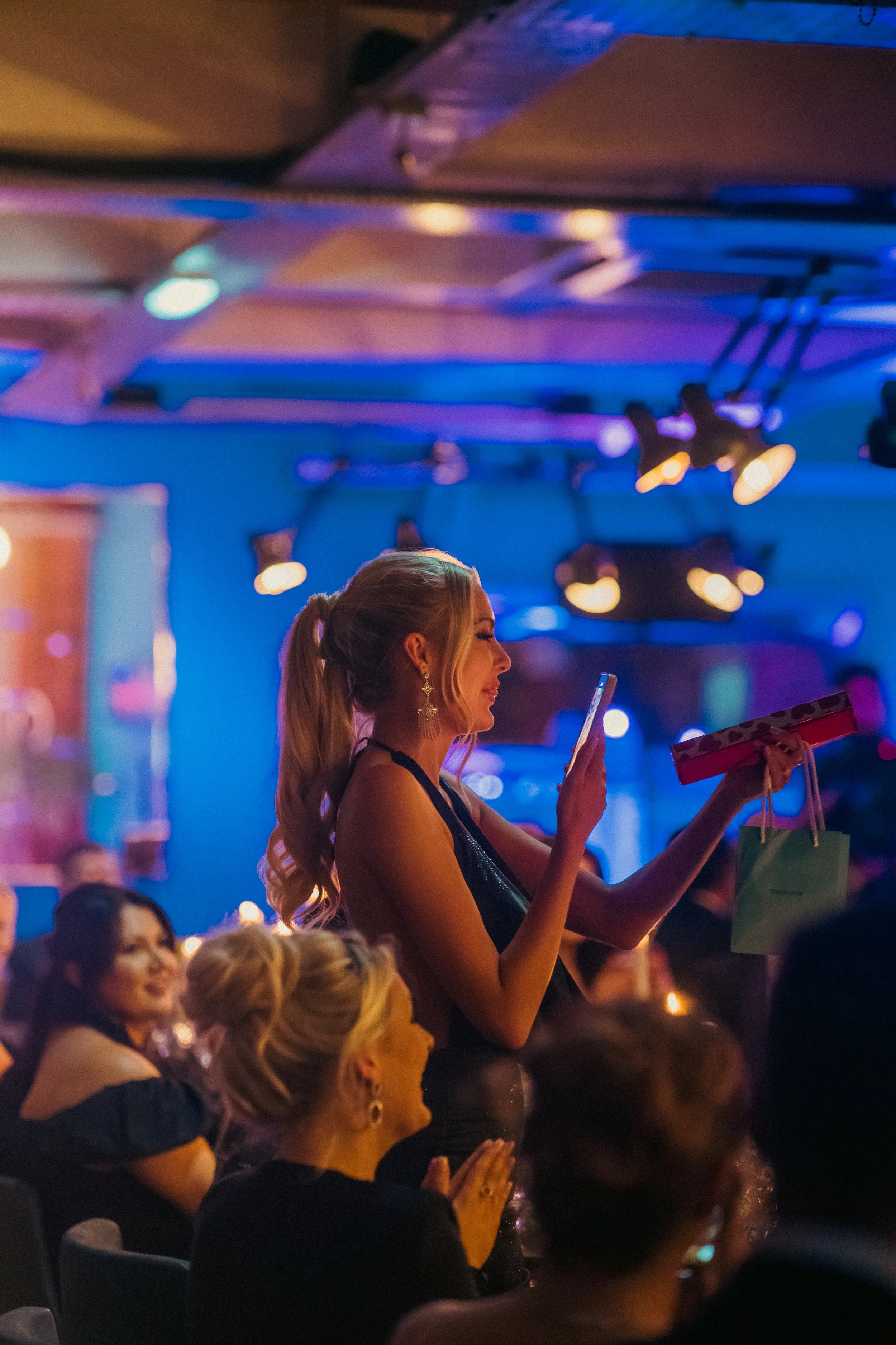 A woman in a black dress with earrings holding a gift and a shopping bag, smiling while a woman behind her looks on in a colorful, dimly lit setting with people seated at tables.