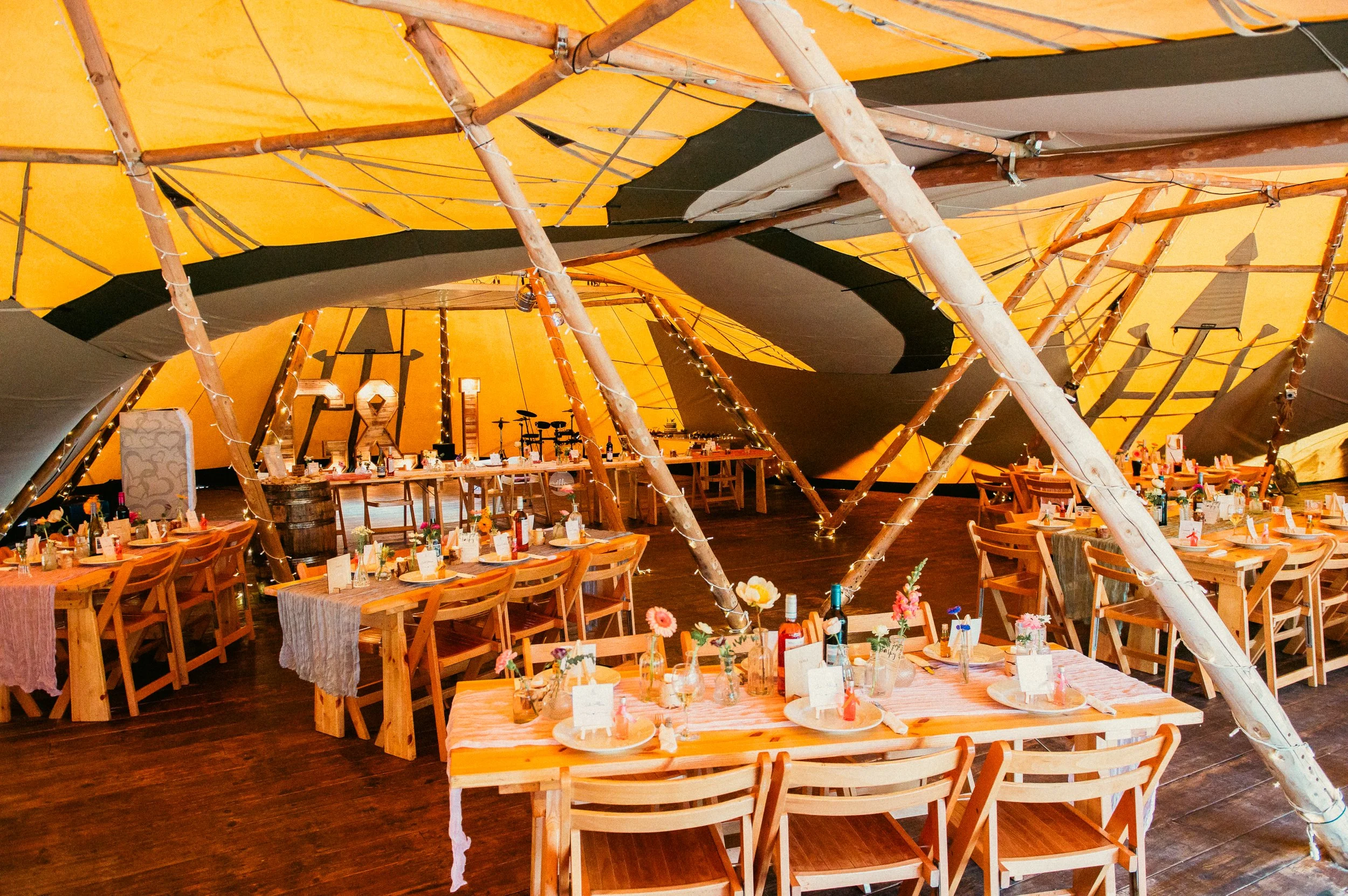 Interior of a decorated tent with multiple tables set for a celebration, adorned with flowers, bottles, and place settings, under a large yellow and black canopy, with a stage area in the background.