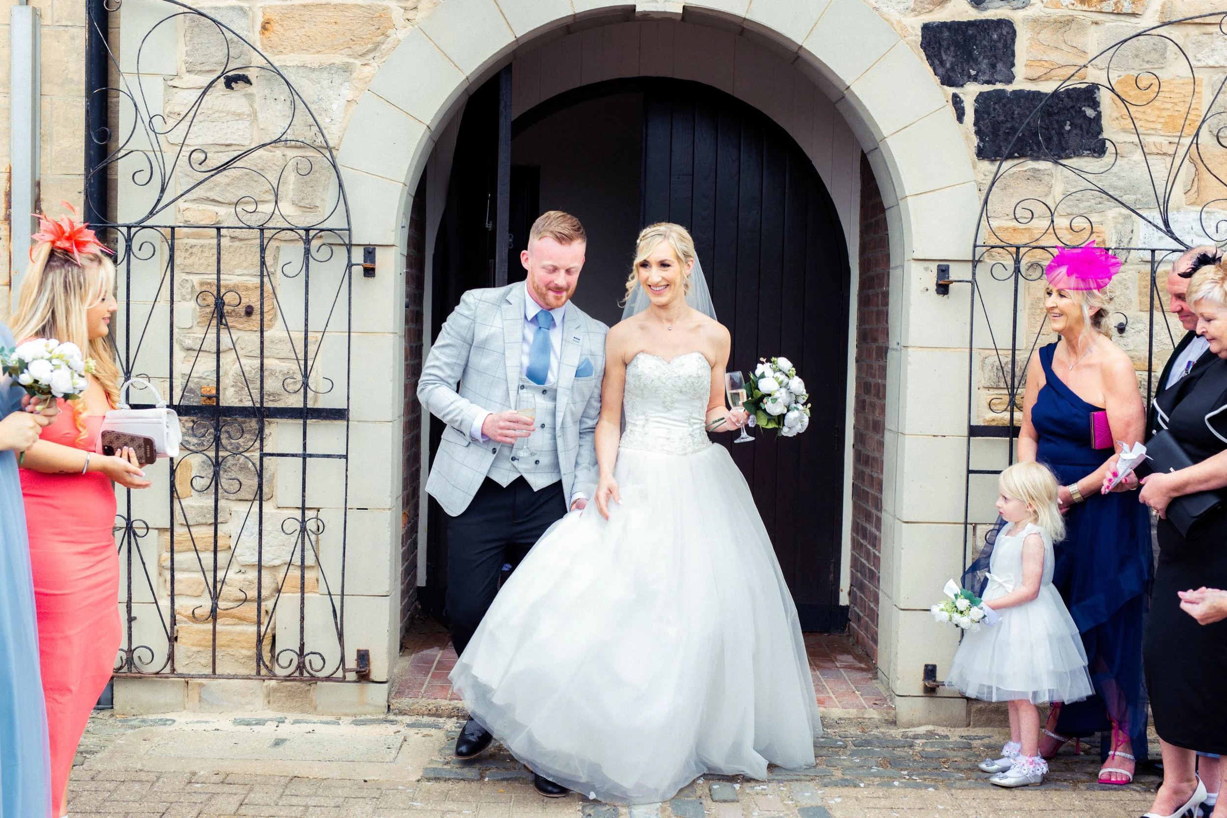 A bride and groom exit a church or wedding venue, surrounded by guests. The bride is wearing a white strapless wedding gown and holding a bouquet of white flowers. The groom is dressed in a light gray checked suit with a light blue shirt and tie. Gue