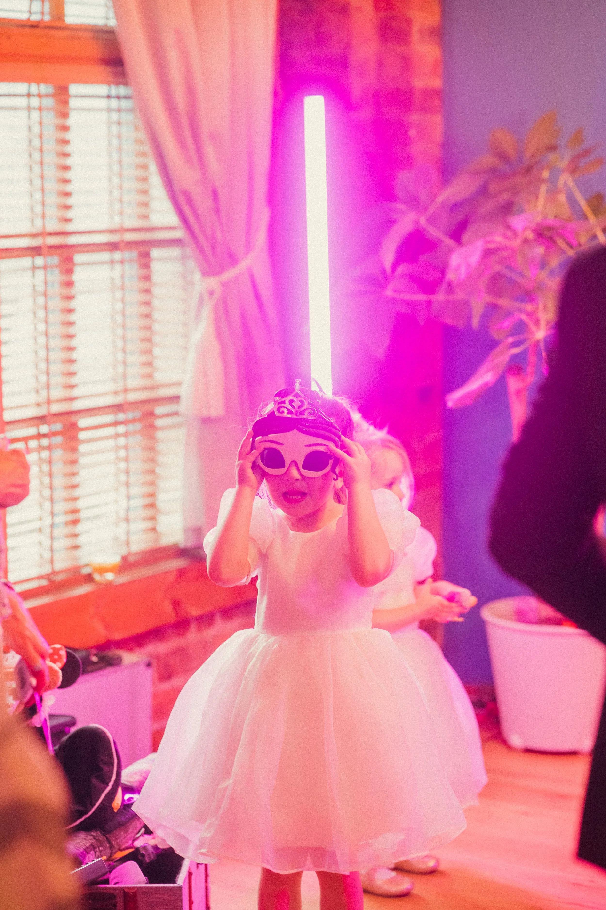 A young girl in a white dress wears oversized sunglasses and a tiara, adjusting her sunglasses at a birthday party with colorful lighting and decorations.