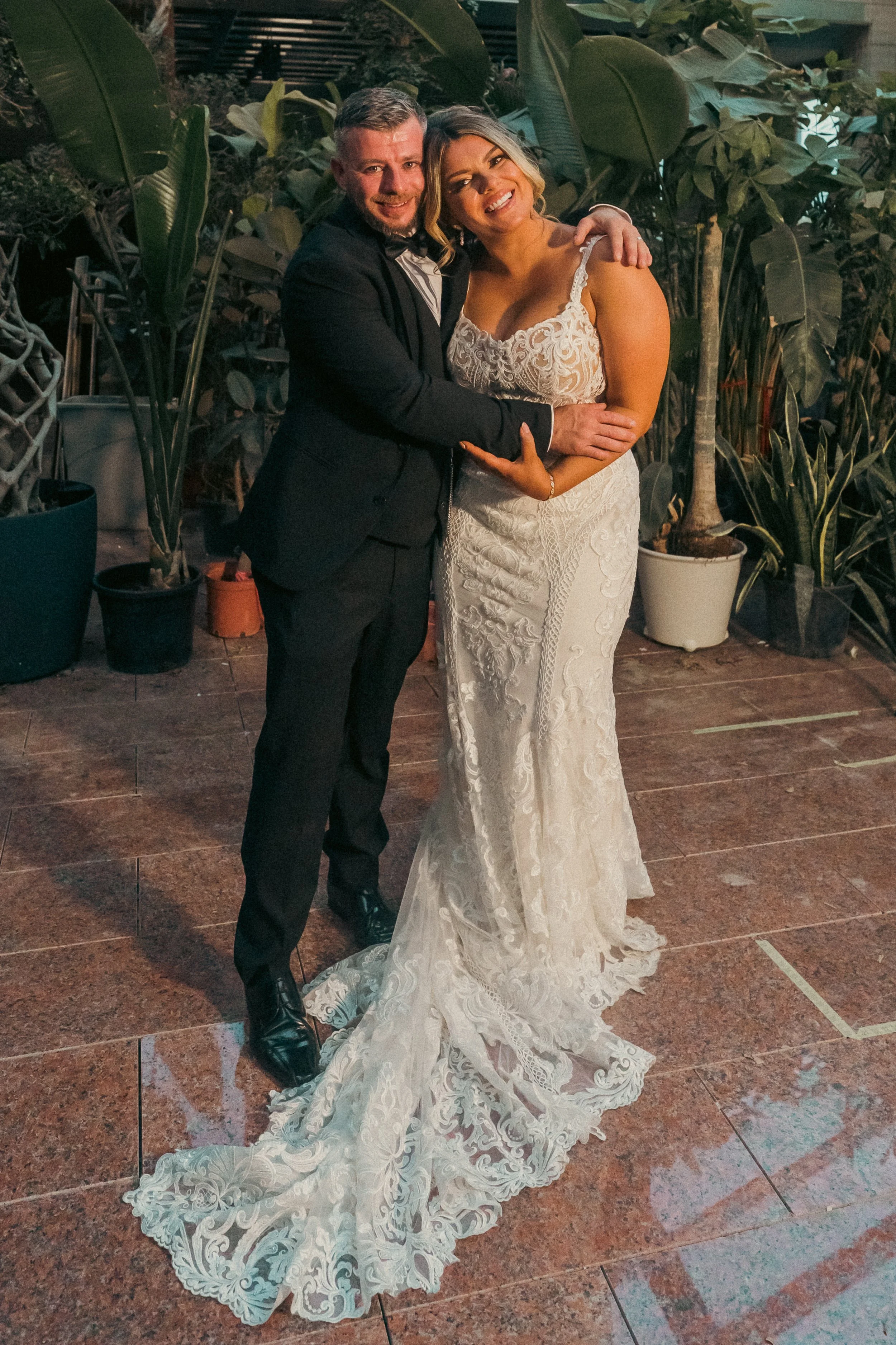A couple in wedding attire embracing and smiling at a social event, with plants in the background.
