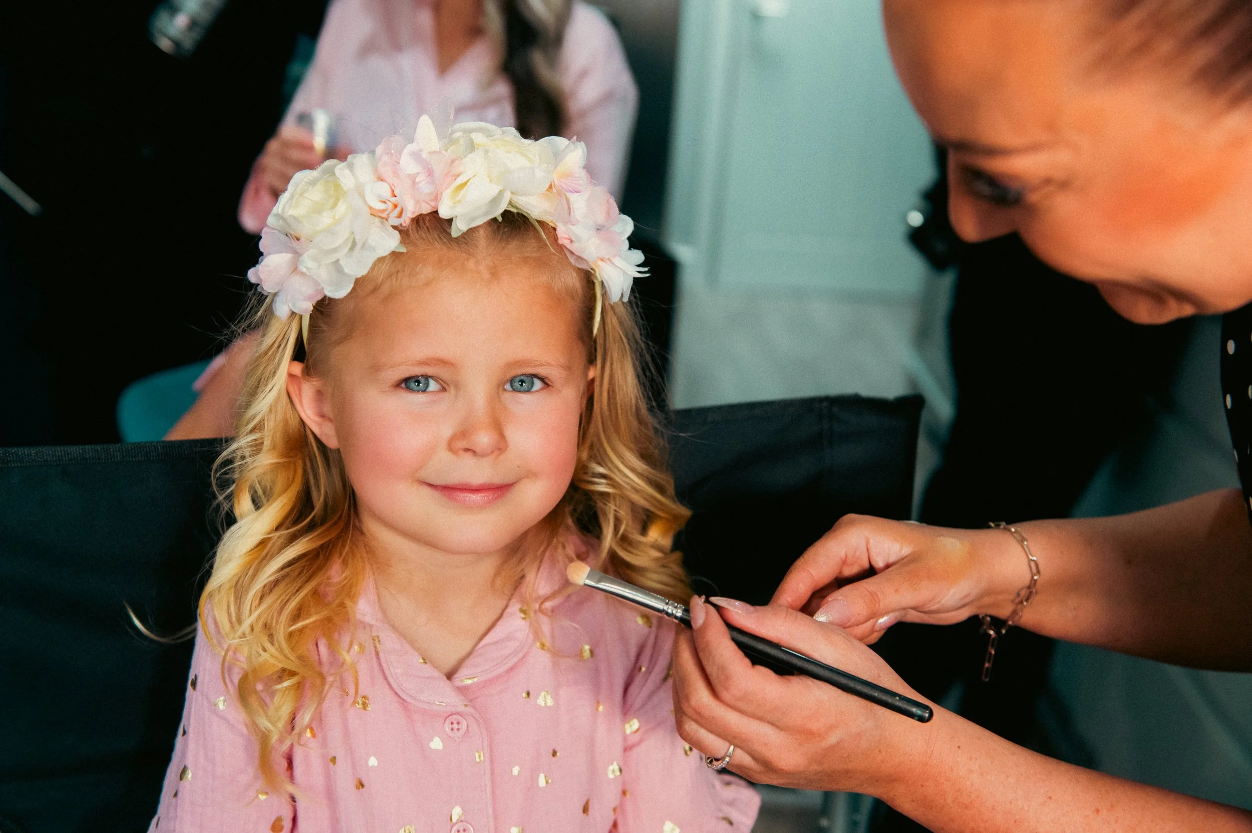 A young girl with blonde hair and blue eyes, wearing a pink shirt with gold heart patterns and a flower crown, gets her face painted by a woman.
