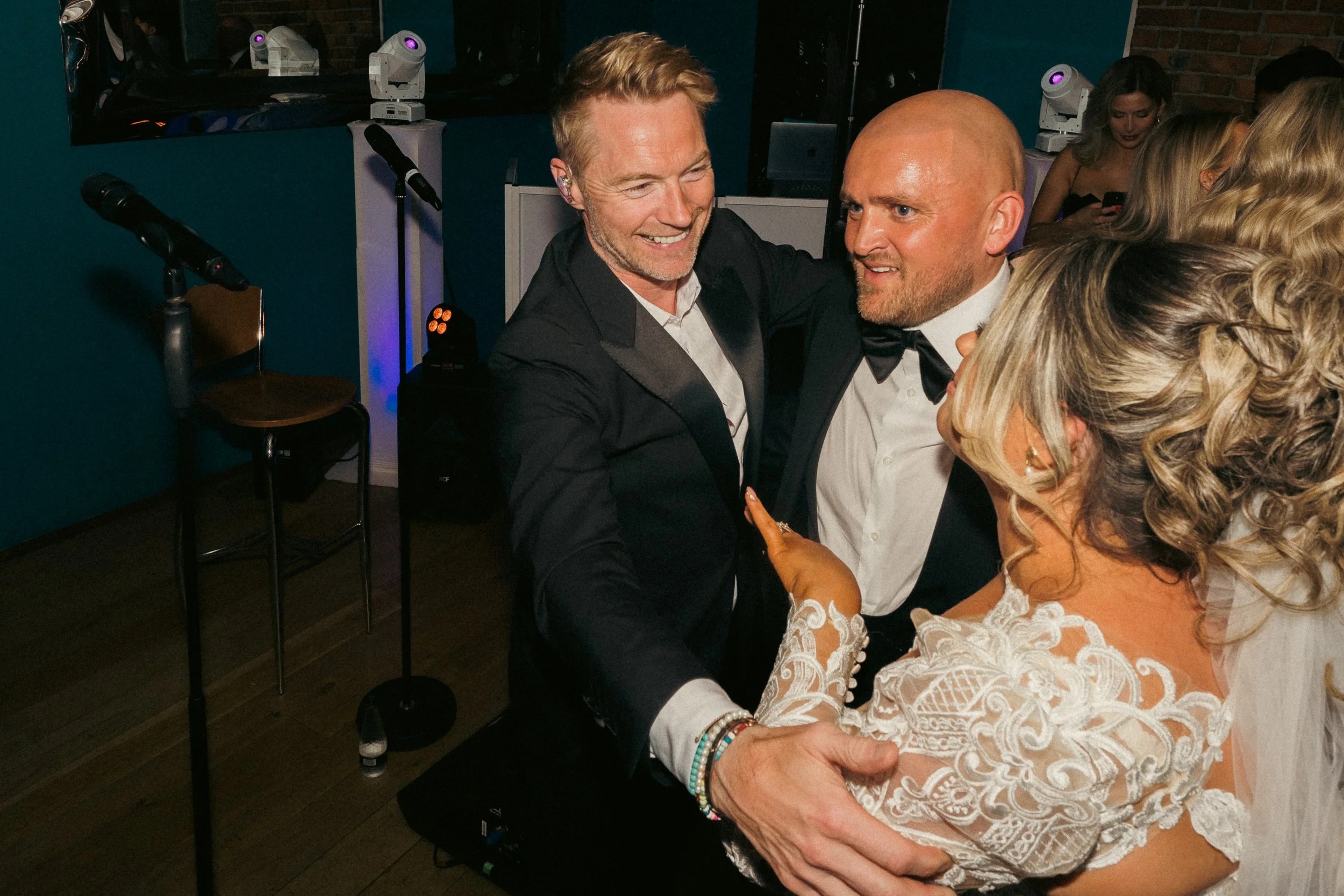 People dancing at a wedding reception, including a woman in a lace dress, a man with a shaved head and bow tie, and another man in a tuxedo, all smiling and enjoying the celebration.