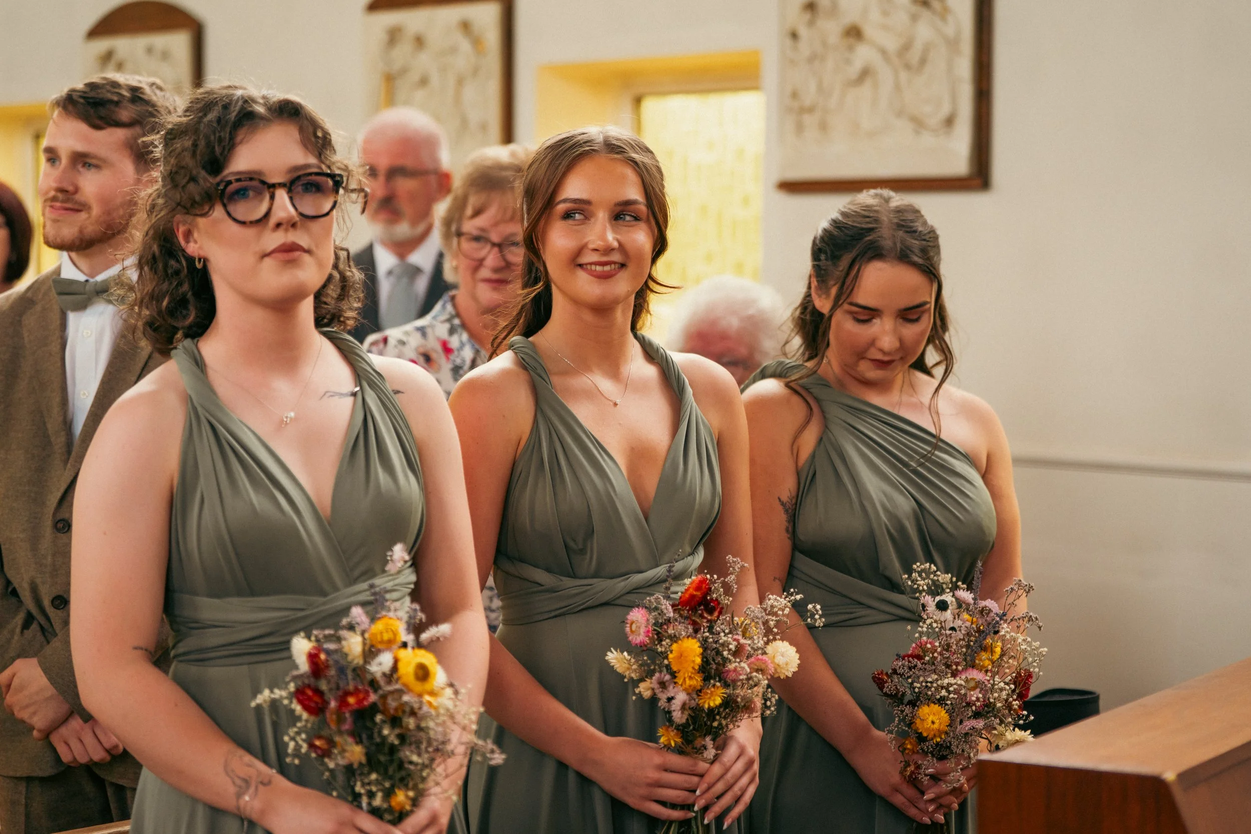 Three women in matching gray dresses holding bouquets of flowers, standing at a wedding ceremony inside a church.
