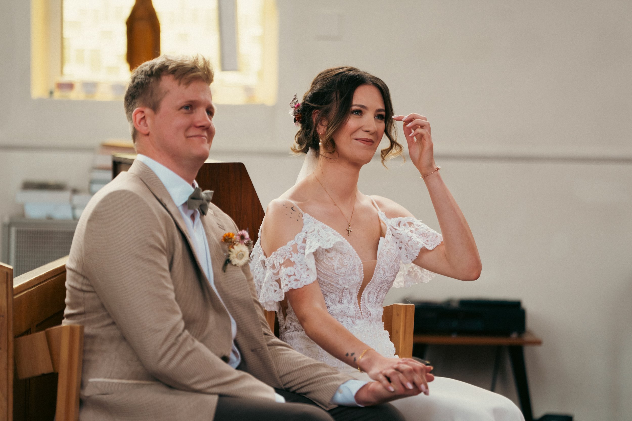 A bride and groom sitting in a church, holding hands during their wedding ceremony. The bride has dark hair styled in loose curls and is wearing a white lace wedding dress, while the groom has light hair and is dressed in a tan suit with a bow tie.
