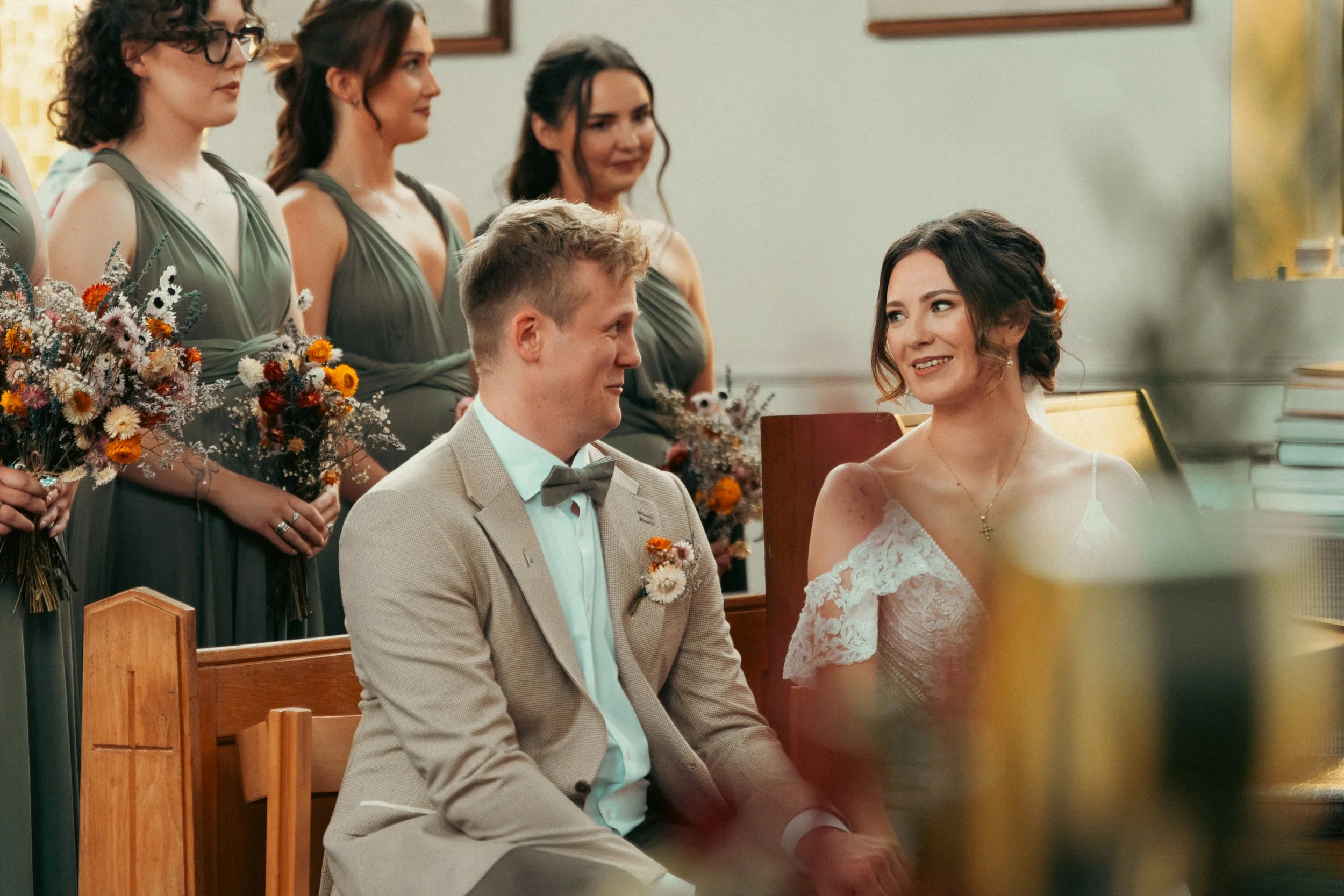 A wedding ceremony with a bride and groom sitting together, smiling at each other, and four bridesmaids standing behind them holding bouquets of flowers.