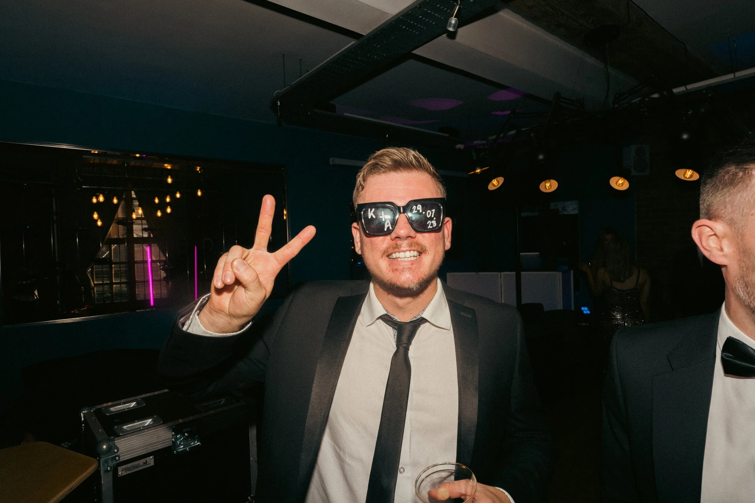 A smiling man in a tuxedo and sunglasses making a peace sign at a party or celebration event.