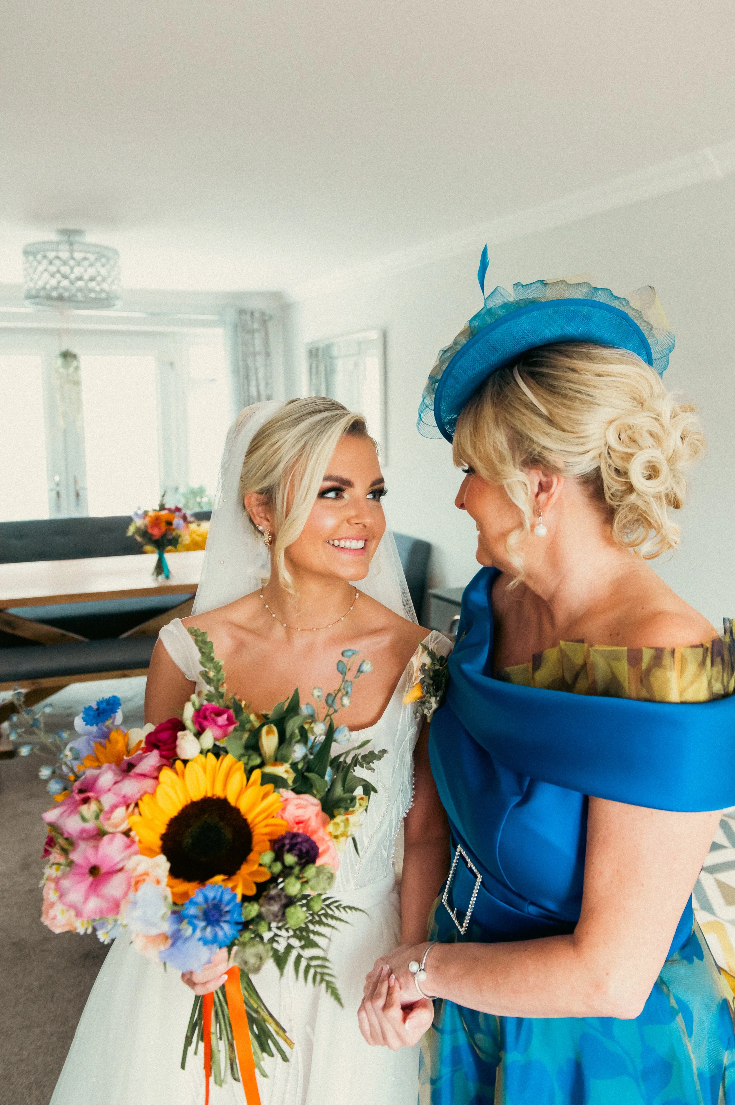 Bride in a white wedding dress and veil holding a colorful bouquet, smiling at a woman in a vibrant blue dress and matching hat in a bright indoor setting.