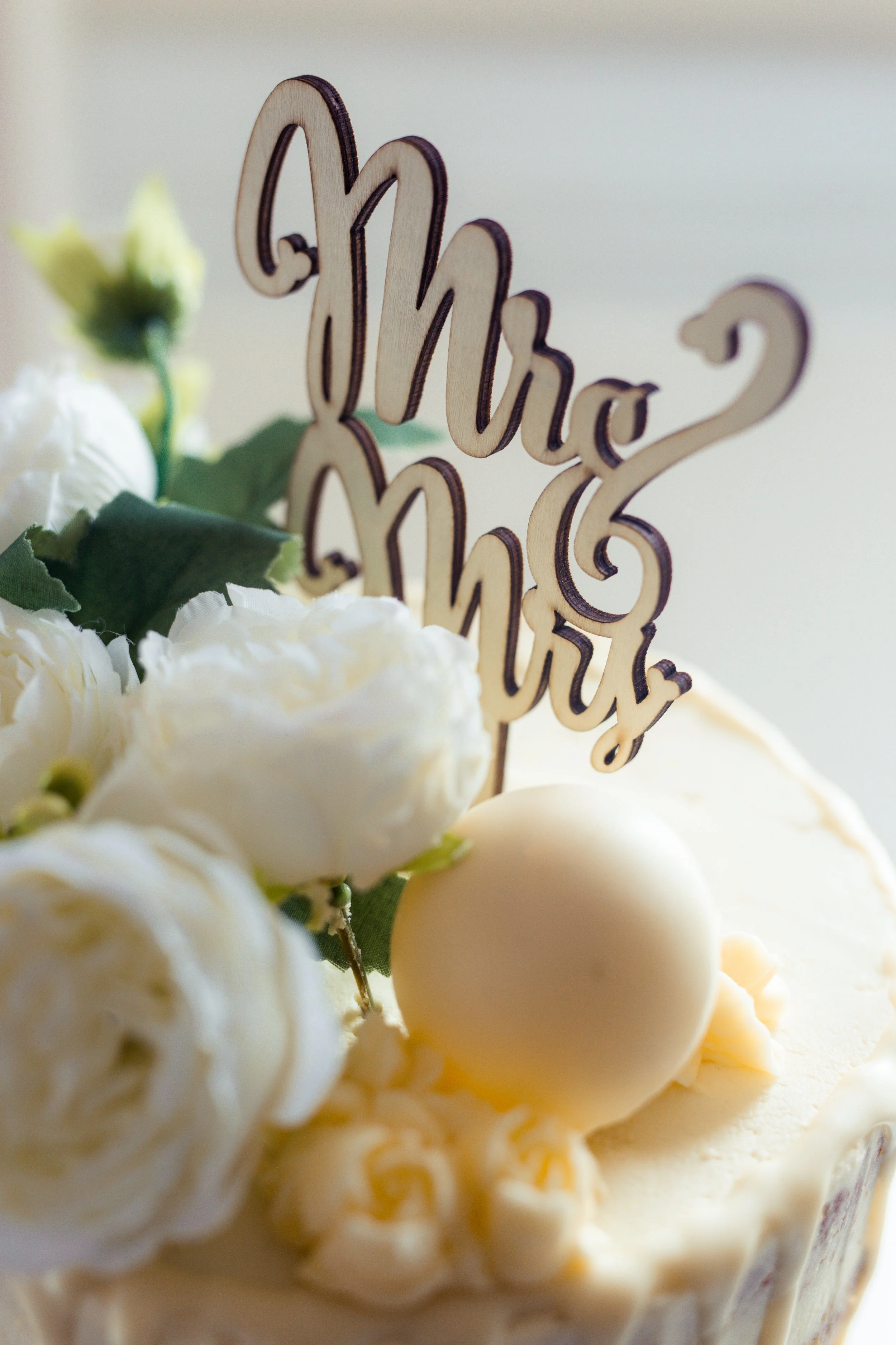 A cake decorated with white flowers, a round white ornament, and a wooden 'Mrs & Mrs' topper.