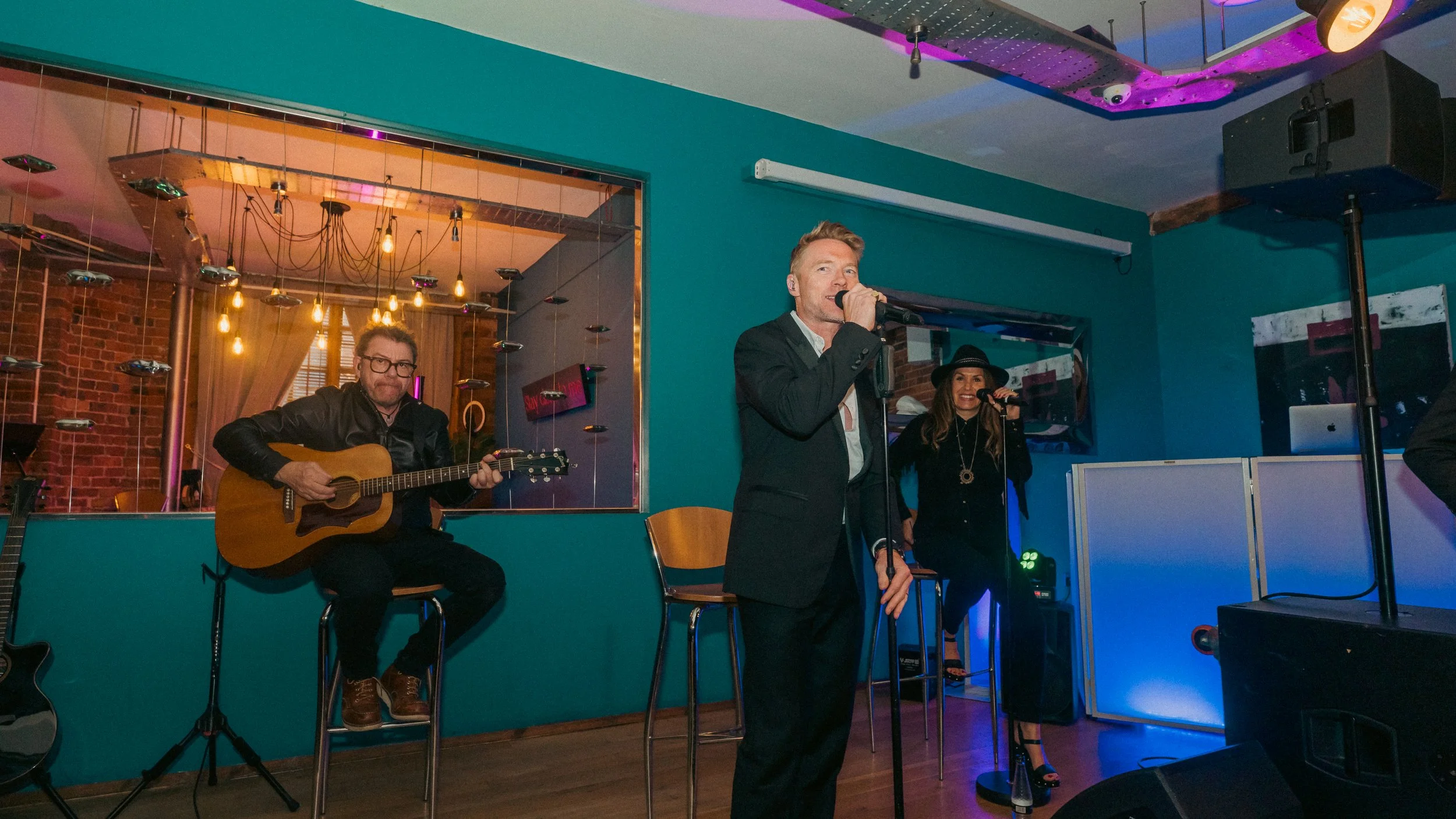 A band performs at a cozy indoor venue with teal walls, featuring a male singer in a dark suit and two backup singers, one playing guitar and another singing, all under warm stage lighting.
