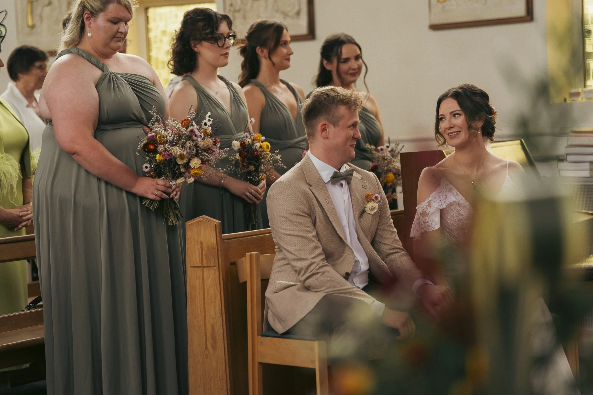 A wedding ceremony inside a church, with a bride and groom seated, smiling at each other, surrounded by bridesmaids holding bouquets of flowers, and guests in the background.