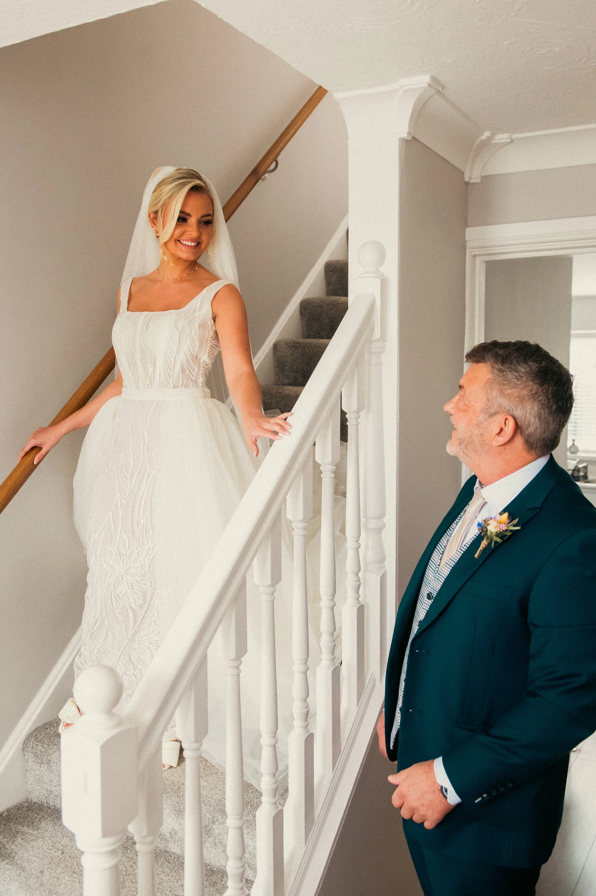 A bride in a white wedding dress and veil smiling while looking at a man in a teal suit, who is smiling back at her, inside a home near a staircase.