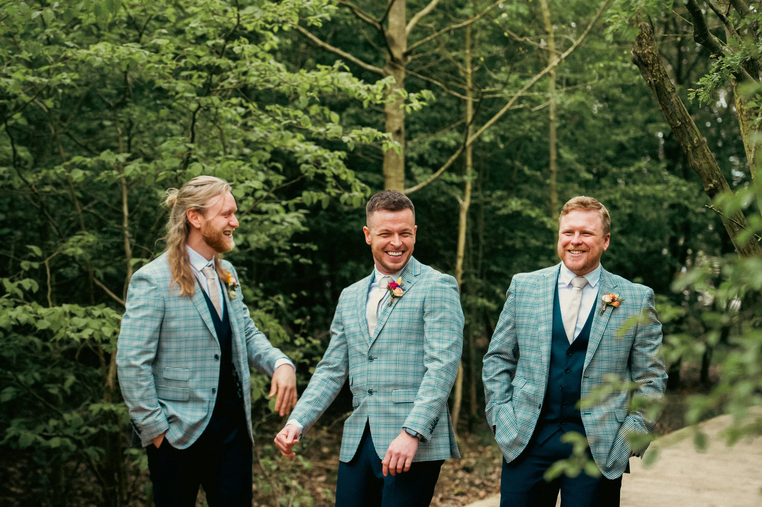 Three men in matching blue plaid suits and white shirts, smiling and walking outdoors in a lush green forest.